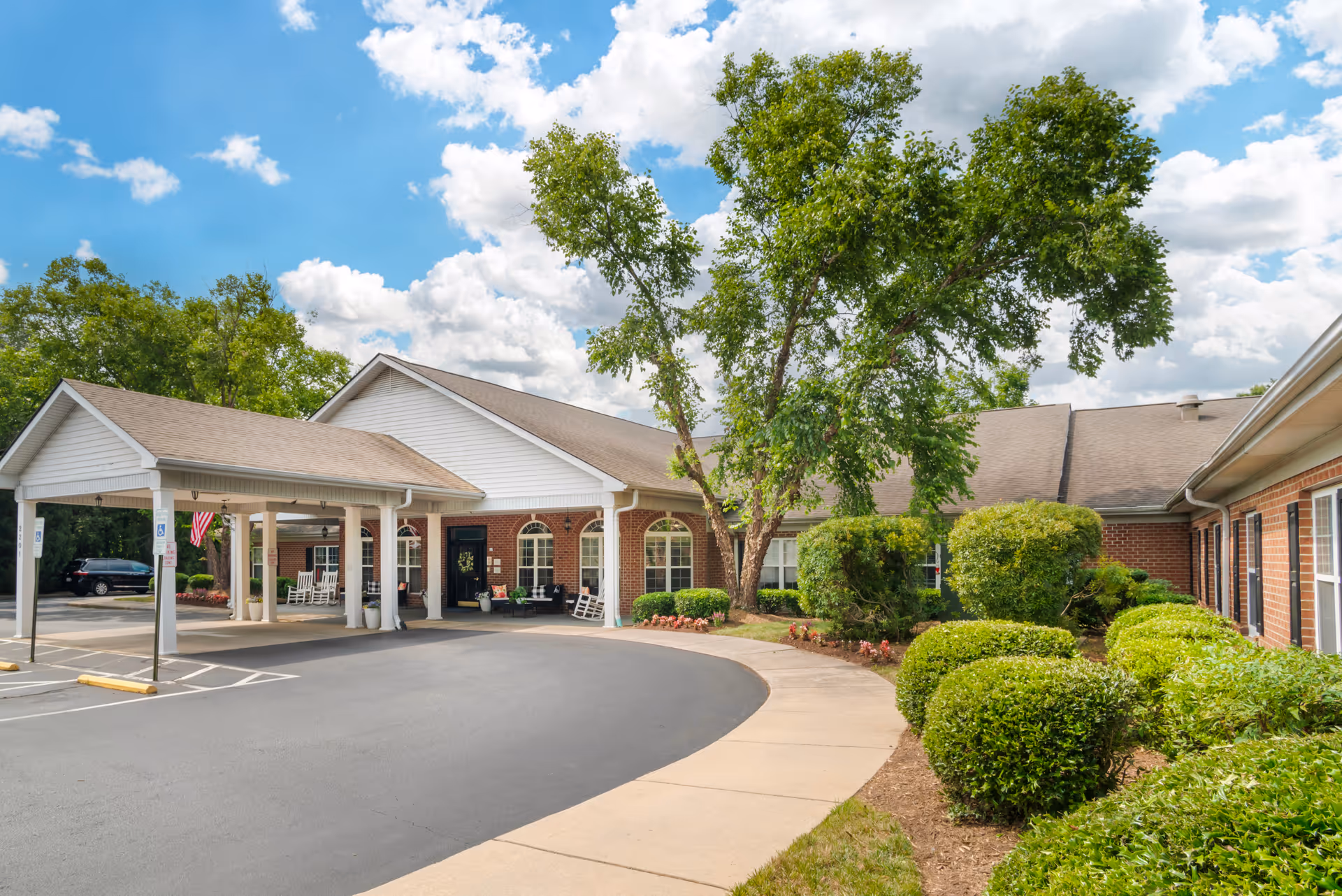 Exterior view of Brookdale Salisbury facility showing a single-story brick building with a covered entrance, rocking chairs on the porch, landscaped bushes, trees, and a clear blue sky with some clouds.