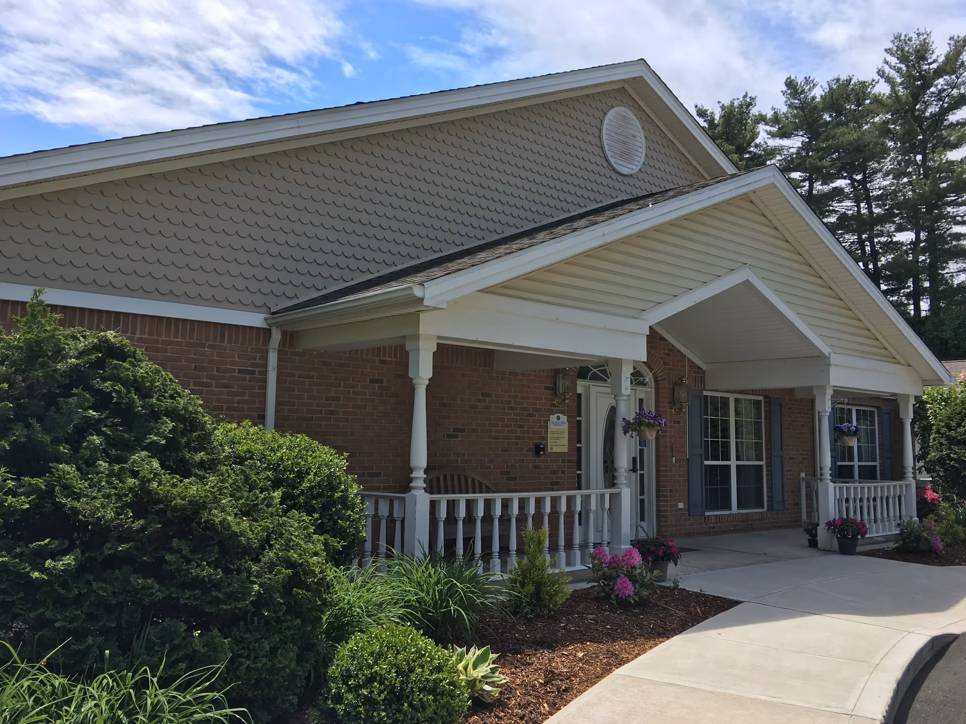 Exterior view of a single-story brick building with a covered porch entrance. The porch has white railings and columns, hanging flower pots with purple flowers, and landscaped bushes and plants along the walkway. The sky is partly cloudy with blue patches visible.
