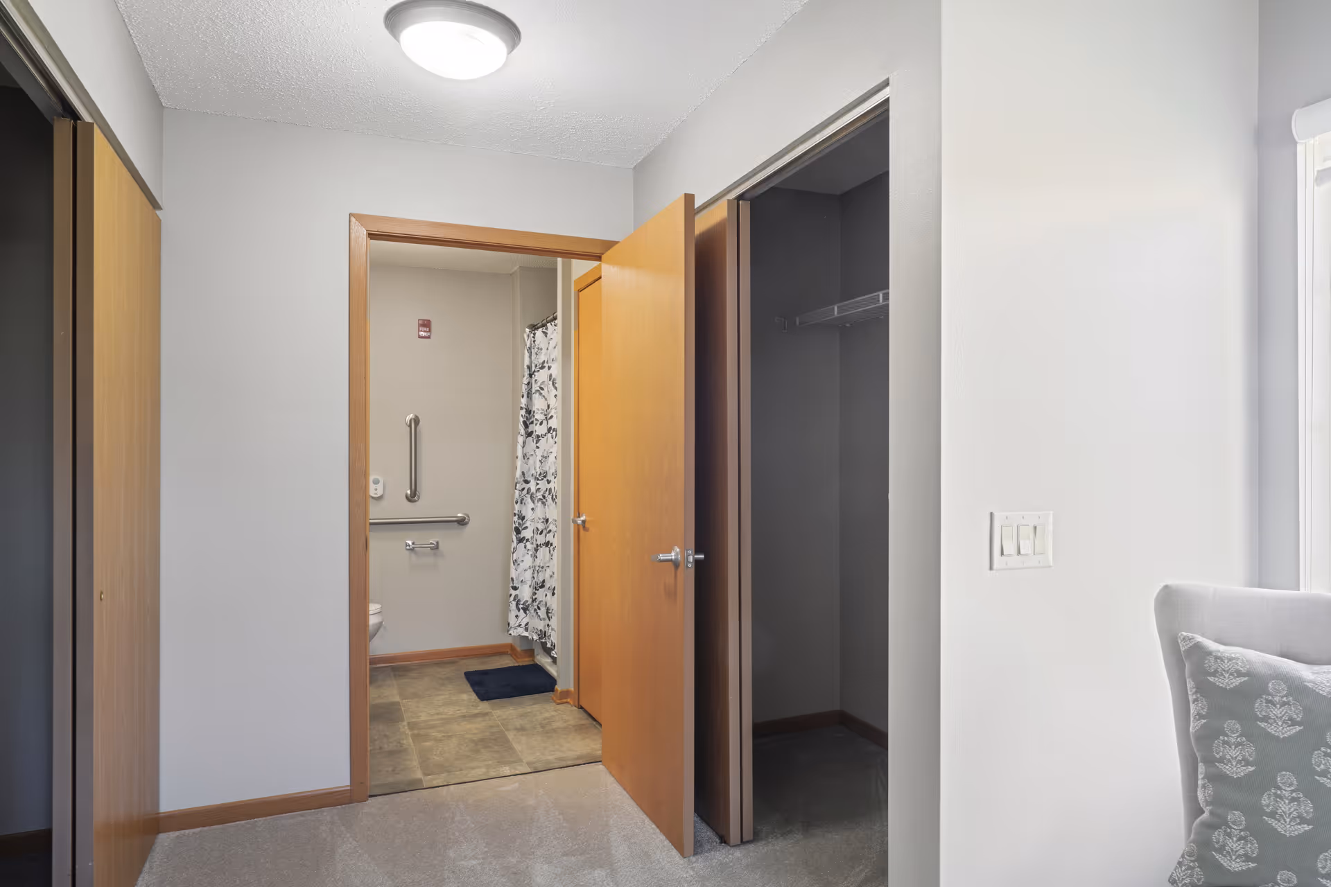 View of a hallway leading to a bathroom with an open door. The bathroom has a grab bar, toilet paper holder, and a shower curtain with a floral pattern. There is a closet with an open door on the right side of the hallway and another closed closet door on the left. Part of a cushioned chair with a patterned pillow is visible on the right side near a window.