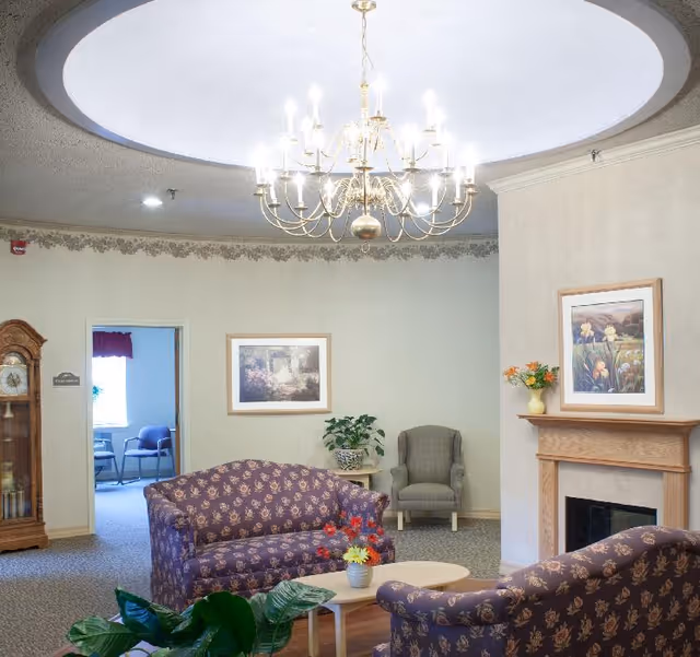 A cozy living room area with two purple patterned sofas facing each other around a small wooden coffee table with a flower vase. There is a gray armchair in the corner next to a small table with a plant. A wooden fireplace with a framed floral painting above it is on the right wall. A large chandelier hangs from a circular recessed ceiling light. A grandfather clock stands near a doorway leading to another room with chairs and a window.