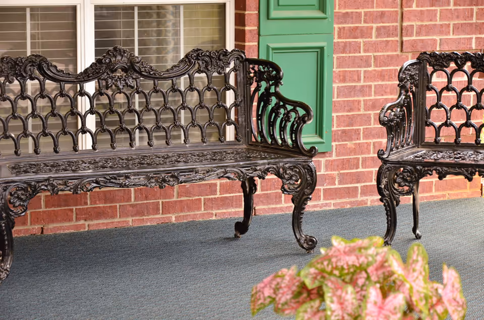 Ornate black metal benches on a covered brick front porch beside a window with a green shutter and a potted plant.