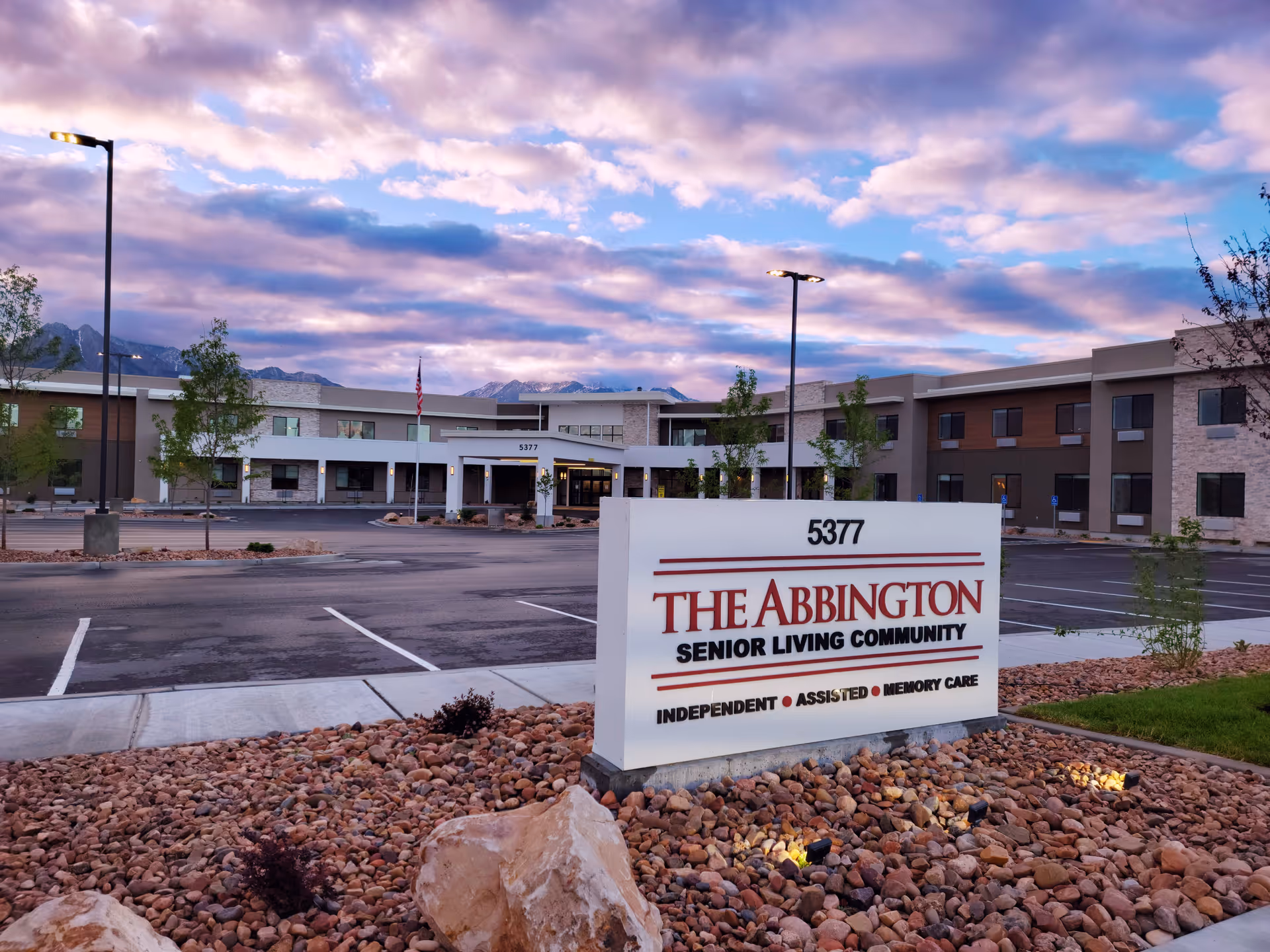 Exterior view of The Abbington Senior Living Community building at dusk with a large sign in the foreground displaying the facility name, address 5377, and services offered: Independent, Assisted, and Memory Care. The building is modern with multiple windows and surrounded by a parking lot, small trees, and landscaping with rocks.