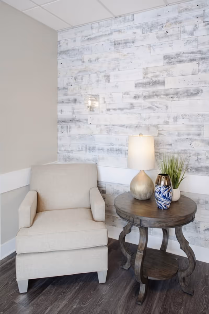 Beige armchair beside a round wooden side table with a lamp and decorative vases against a whitewashed wood-plank accent wall.