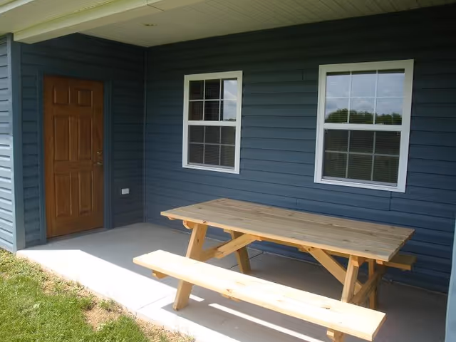 Outdoor covered patio area with a wooden picnic table and bench set on a concrete floor, next to a blue exterior wall with two white-framed windows and a wooden door.