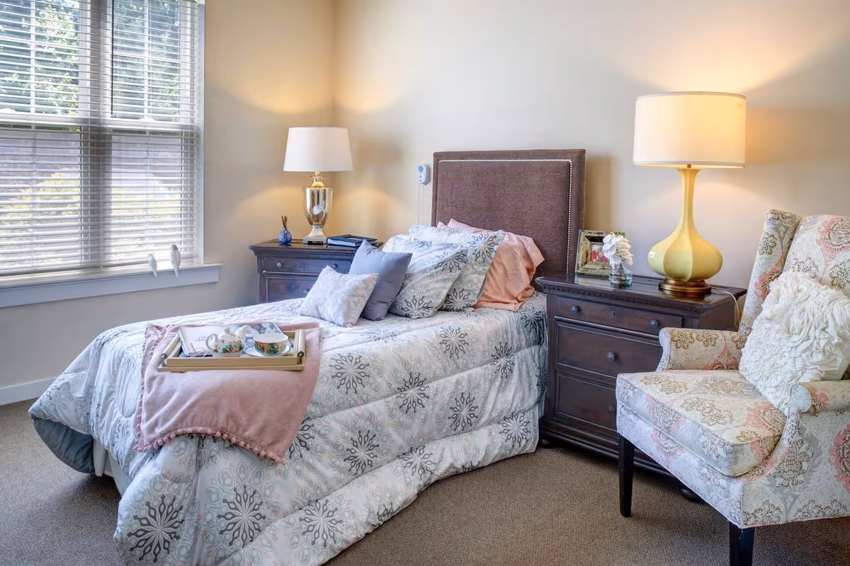 A bright bedroom with a single bed dressed in patterned bedding, two dark wood nightstands with lamps, and an upholstered armchair by a window.