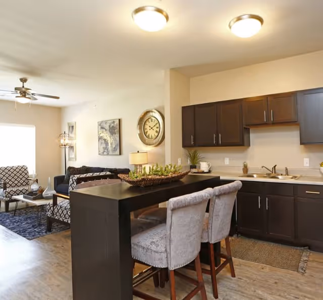 A modern open-concept living area and kitchen in a senior living facility. The kitchen features dark wood cabinets, a double sink, and a black island with two upholstered bar stools. The living area includes patterned armchairs, a dark sofa, a floor lamp, wall art, and a large round clock on the wall. The space has wood flooring and is well-lit with ceiling lights and natural light from a window.