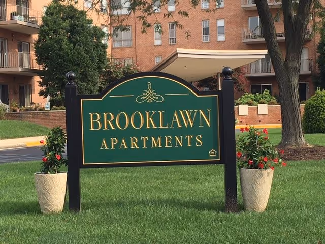 A green and gold sign reading 'Brooklawn Apartments' is displayed on a grassy area with two potted plants with red flowers on either side. In the background, there is a multi-story brick apartment building with balconies and windows.