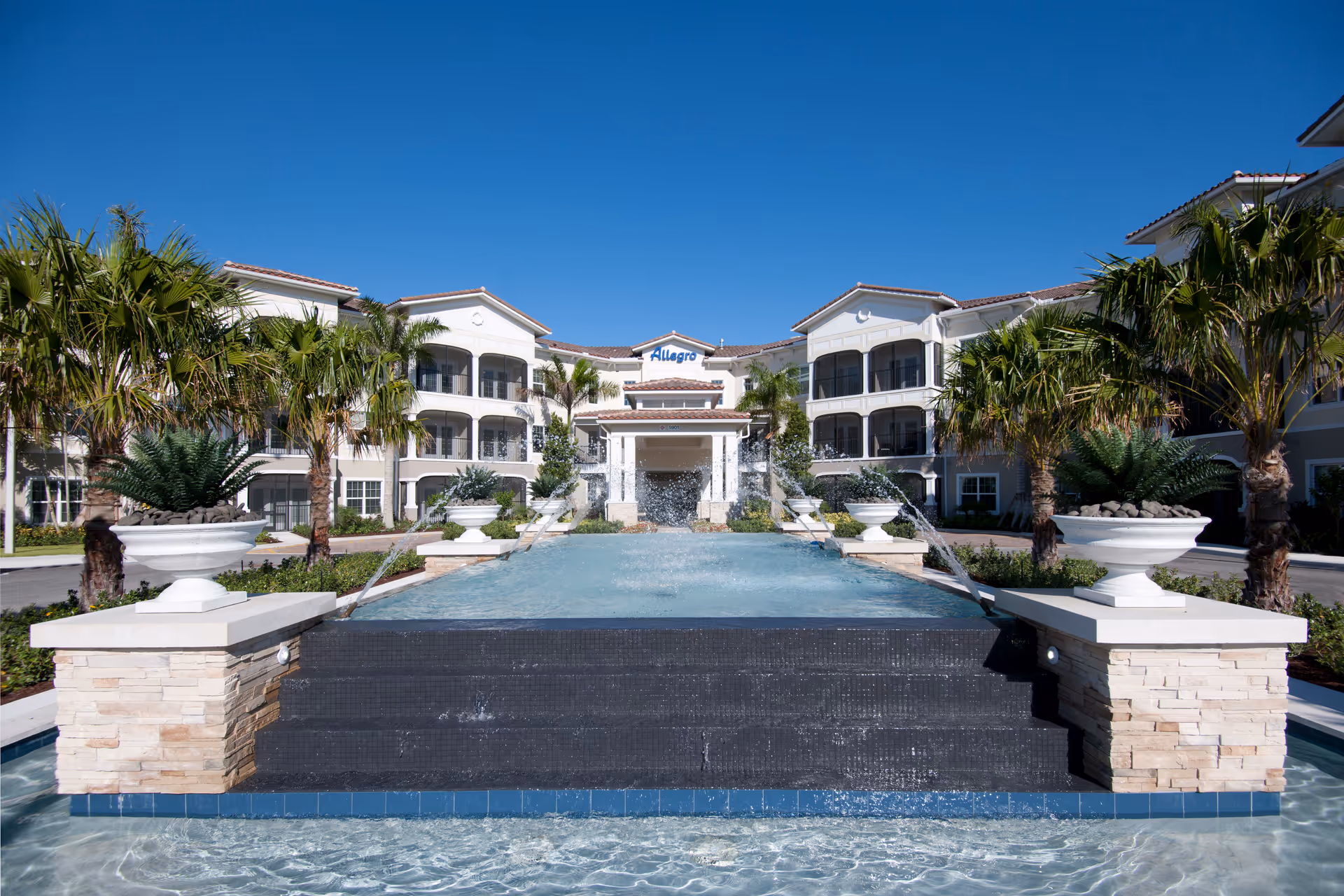 Front exterior view of Allegro Senior Living facility in Parkland, FL, featuring a large water fountain with cascading water, palm trees, and a clear blue sky.