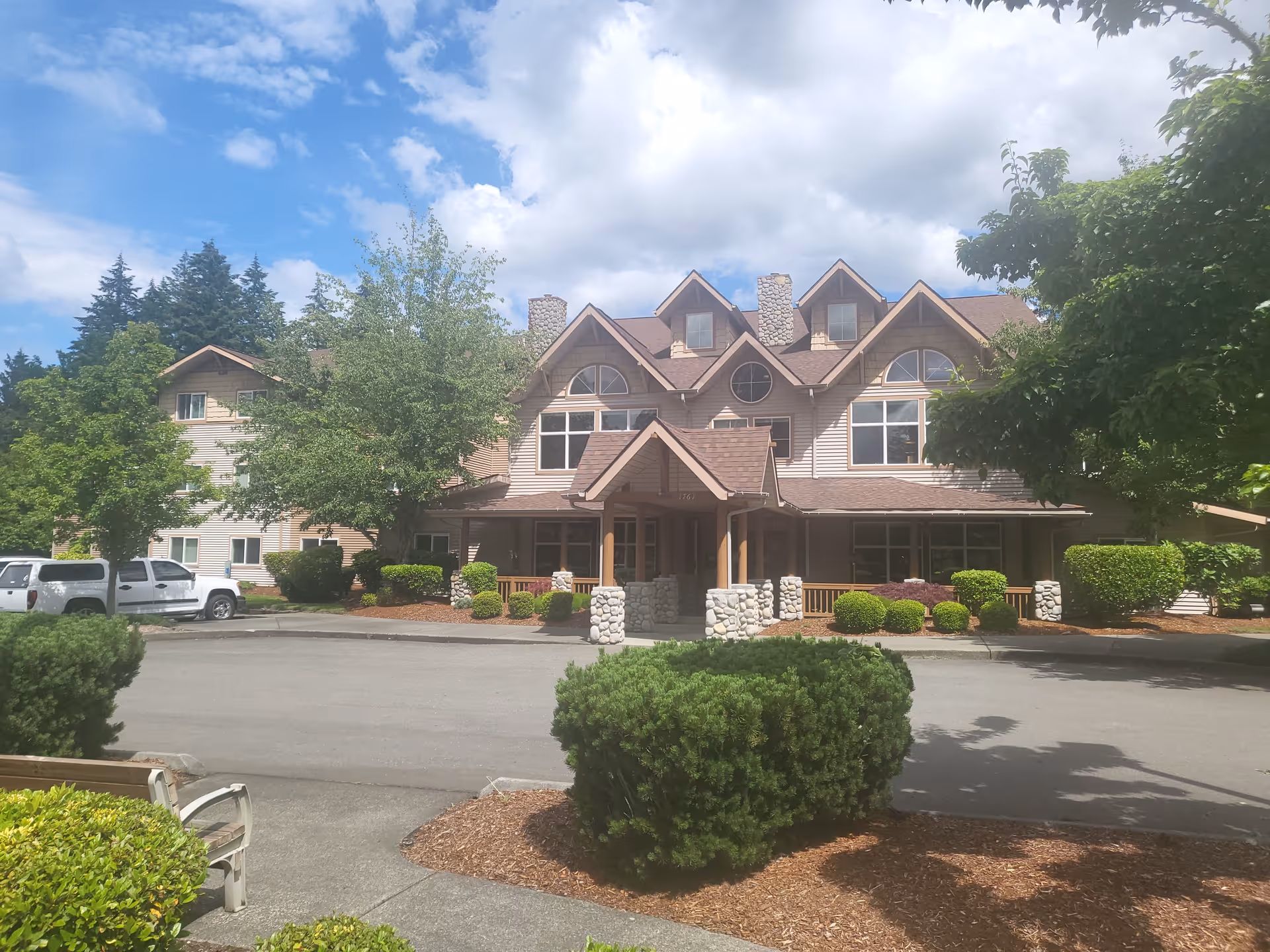 Front exterior view of a multi-story senior living facility building with a peaked roof, stone pillars at the entrance, surrounded by trees, bushes, and a parking area with vehicles under a partly cloudy sky.
