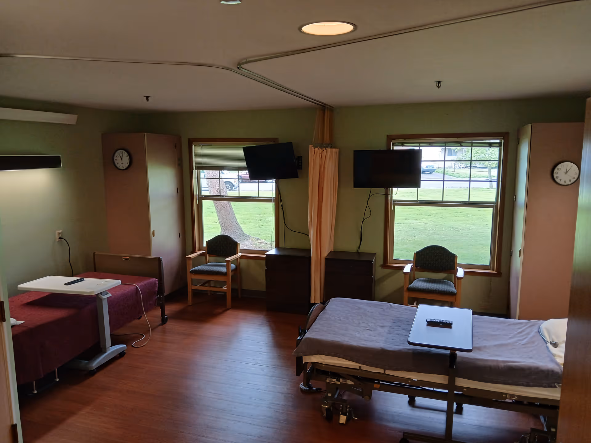 A shared patient room in a healthcare facility with two hospital beds, each with an overbed table and a remote control. The room has two windows with views of green grass outside, two chairs, two dressers, and two clocks on the walls. A privacy curtain is partially drawn between the beds. The walls are painted light green and the floor is wood.