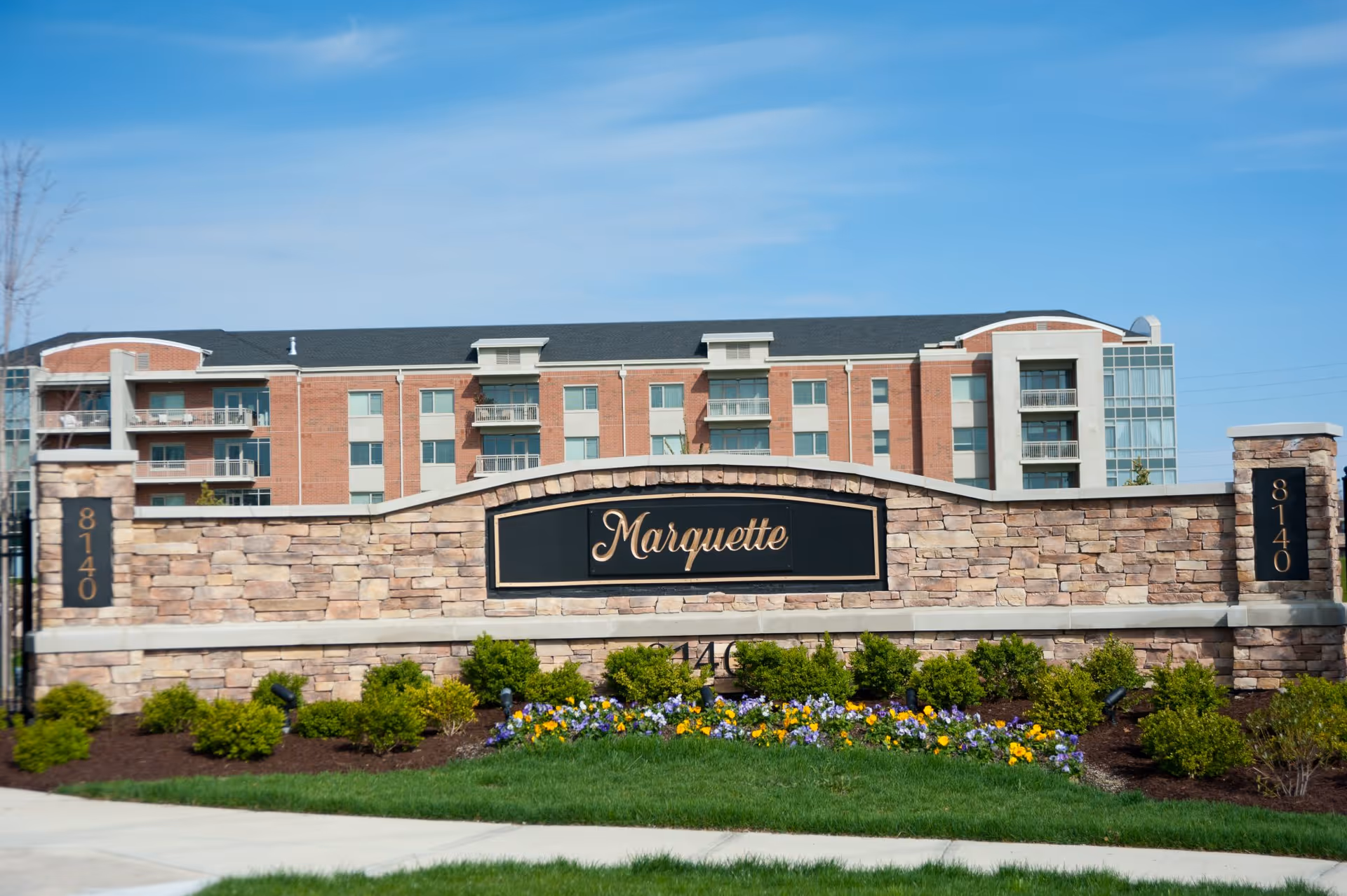 Stone entrance sign reading "Marquette" with landscaped flowers and the multi-story building behind it under a blue sky.