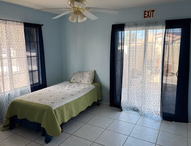 Small bedroom with a single bed, tiled floor, ceiling fan, and a curtained sliding glass door topped by an EXIT sign.