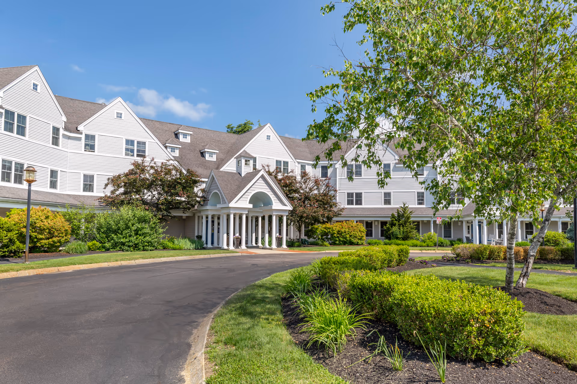 Exterior view of a large senior living facility building with white siding and multiple windows. The entrance features a covered portico with white columns. The surrounding area includes a curved driveway, well-maintained green bushes, trees, and a clear blue sky.
