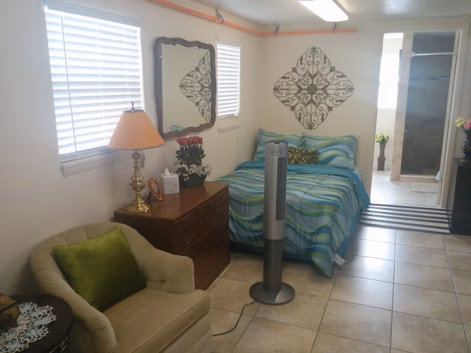 A small bedroom with a bed covered in blue and green striped bedding, a wooden dresser with a lamp, flowers, and a tissue box, a beige armchair with a green pillow, a tall fan in front of the bed, two windows with blinds, a decorative wall mirror, and a metal wall art piece. The room has tiled floors and an open doorway leading to a bathroom with a shower and a flower vase.