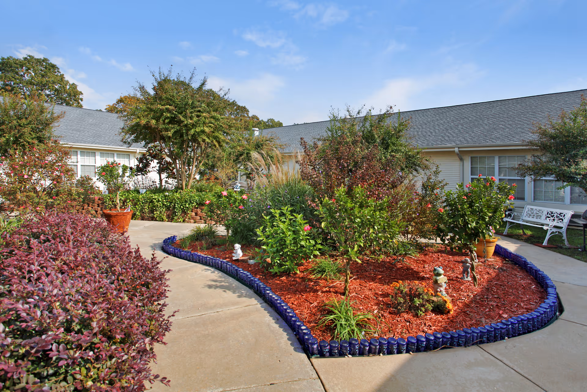 A landscaped garden area with various shrubs, small trees, and flowering plants bordered by blue decorative edging. The garden is surrounded by a concrete walkway and is situated in front of a single-story building with white siding and multiple windows. There is a white bench on the right side near the building. The sky is clear with a few clouds.
