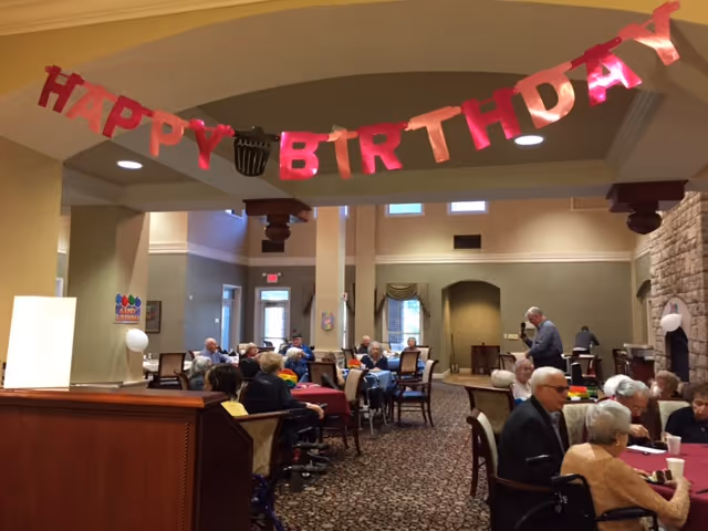 A group of elderly people sitting at tables in a large, carpeted common area decorated with a pink 'HAPPY BIRTHDAY' banner hanging from the ceiling. The room has high ceilings, beige walls, and large windows letting in natural light. Some people are seated in wheelchairs, and others are engaged in conversation.