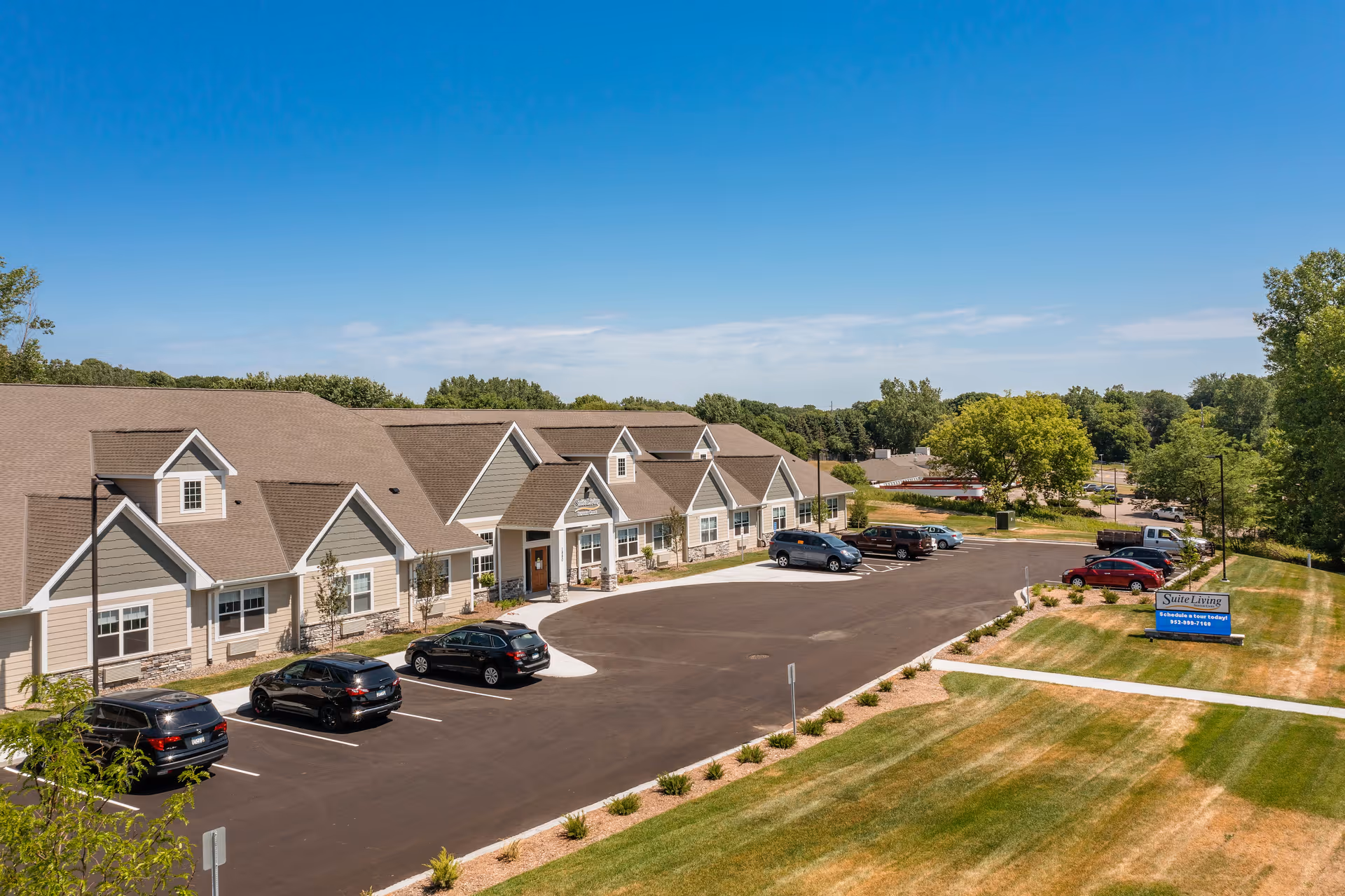 Exterior view of Suite Living of Eden Prairie, a single-story building with a beige and gray facade, multiple gabled roofs, and a parking lot with several cars. The surrounding area includes green lawns, trees, and a clear blue sky.