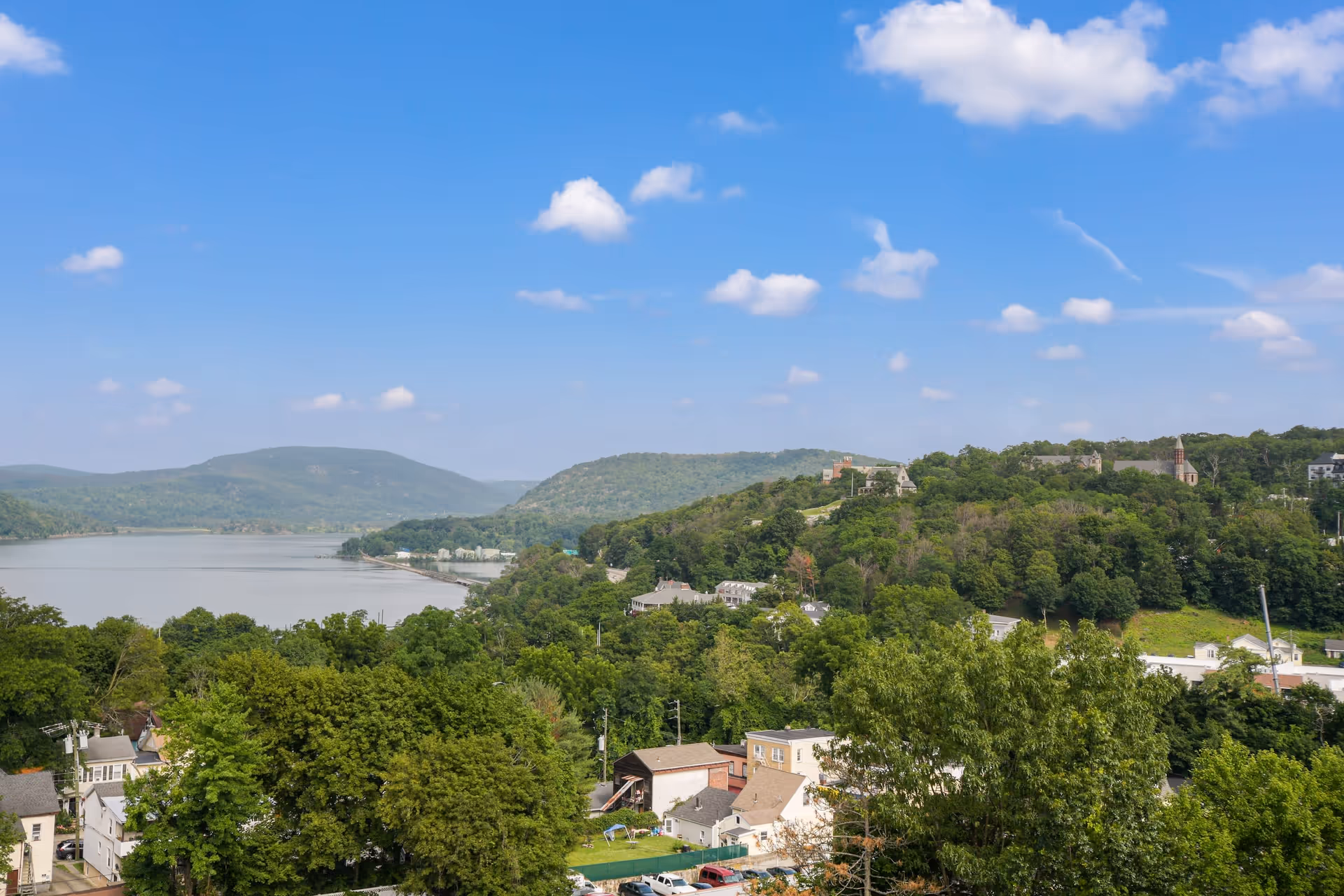 Scenic view of a river with surrounding green hills and trees under a blue sky with scattered clouds. Houses and buildings are visible in the foreground near the riverbank.