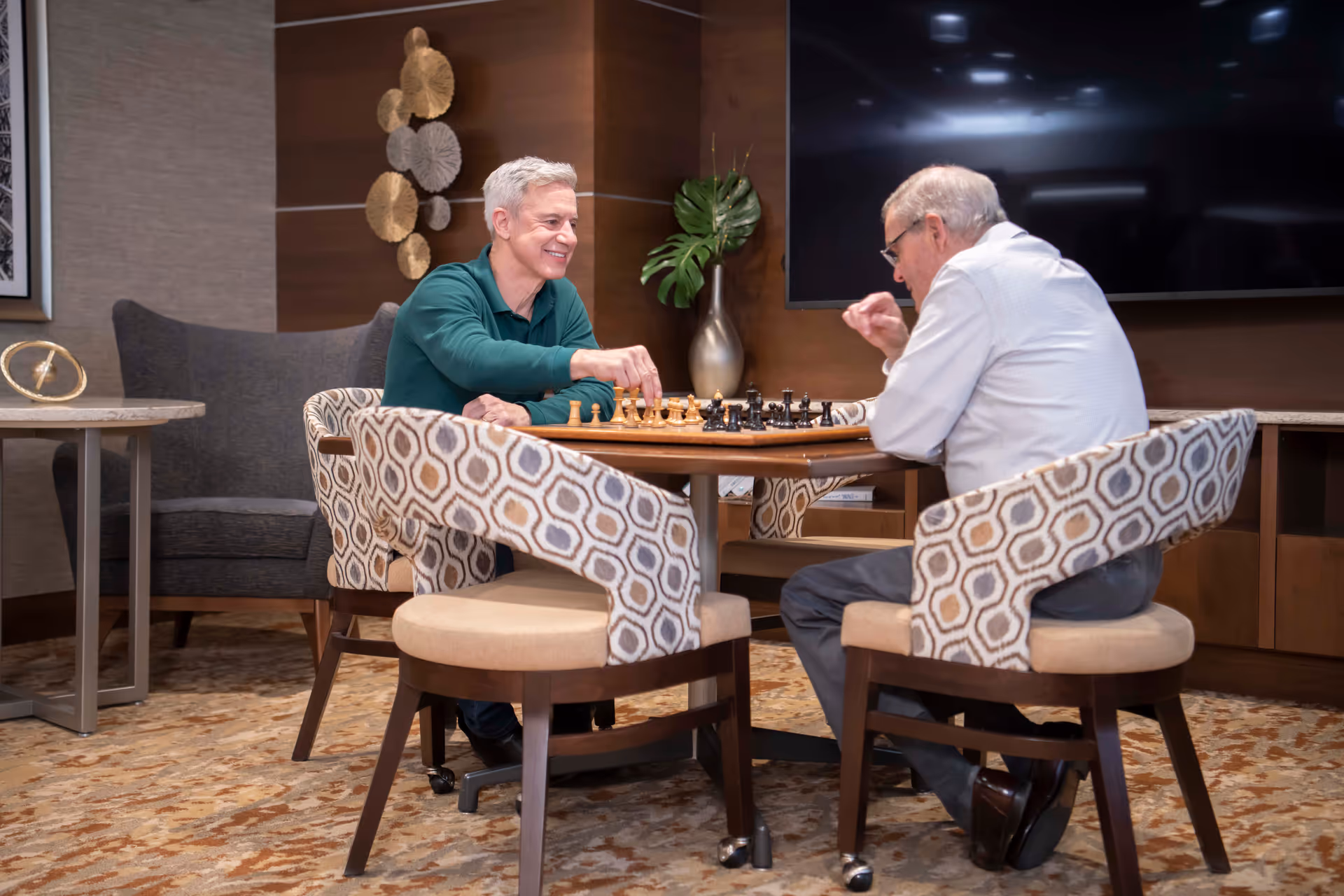 Two elderly men sitting at a round table playing chess in a cozy room with patterned chairs, a large TV mounted on the wall, and decorative elements including a vase with green leaves and wall art.