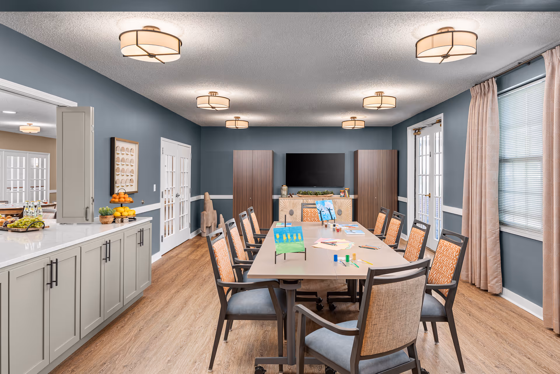 A well-lit activity room with a long table surrounded by chairs. On the table are art supplies and small paintings. The room has blue-gray walls, wood flooring, and multiple ceiling lights. There is a large TV mounted on the far wall between two cabinets, and a countertop with fruit and water bottles on the left side. Windows with beige curtains are on the right side.