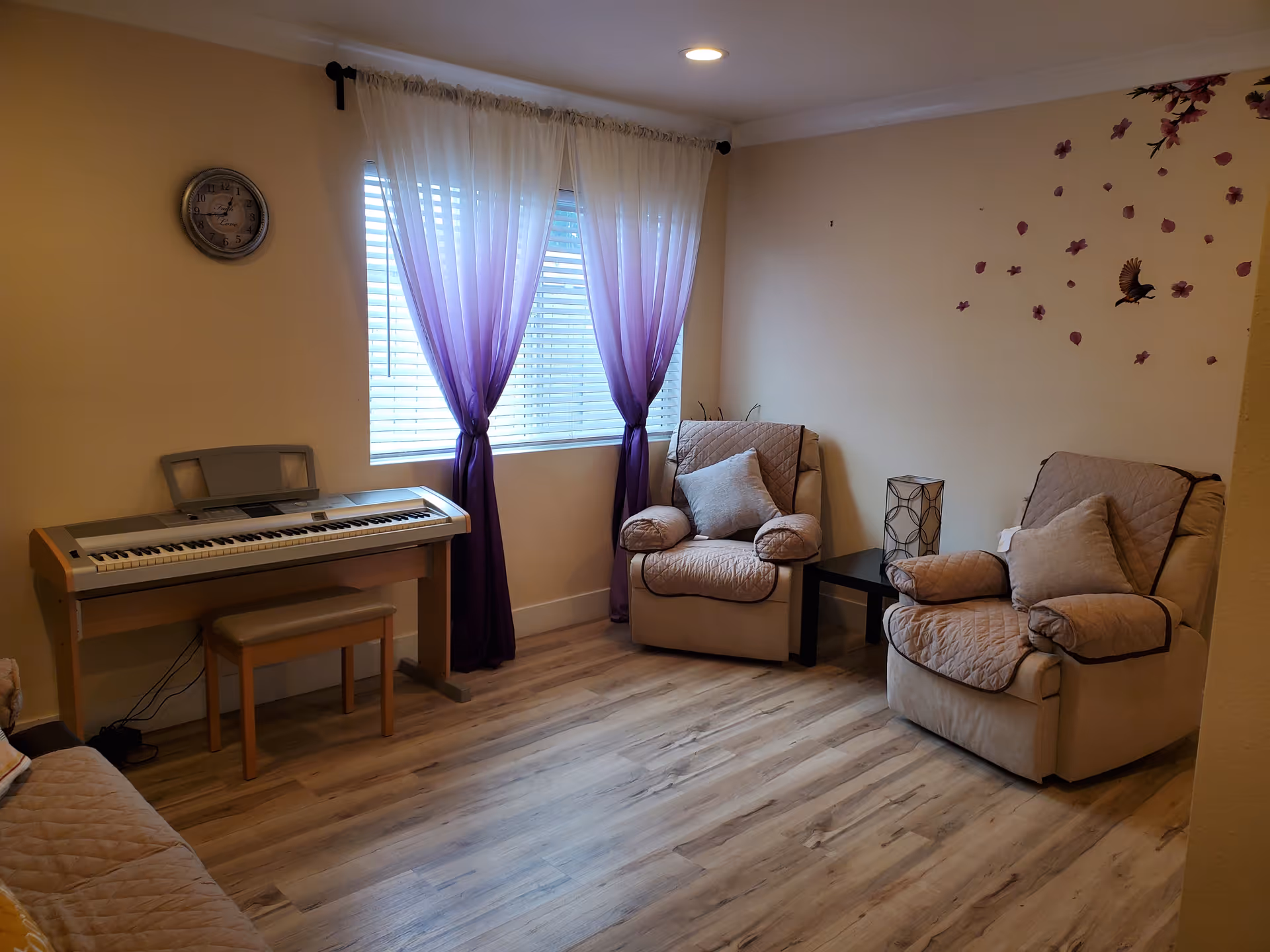 A cozy living room with two beige armchairs covered with quilted cushions, a small black side table with a decorative lamp, a keyboard piano with a bench, and a window with purple ombre curtains letting in natural light. The walls are light-colored with a clock and floral bird wall decals.