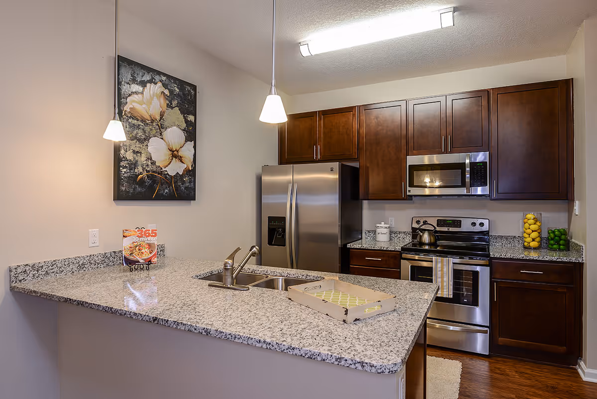 Modern kitchen with granite countertops, stainless steel refrigerator, stove, and microwave. Dark wooden cabinets line the walls. Two pendant lights hang over the countertop with a sink and a decorative tray. A floral painting is mounted on the wall above the counter.
