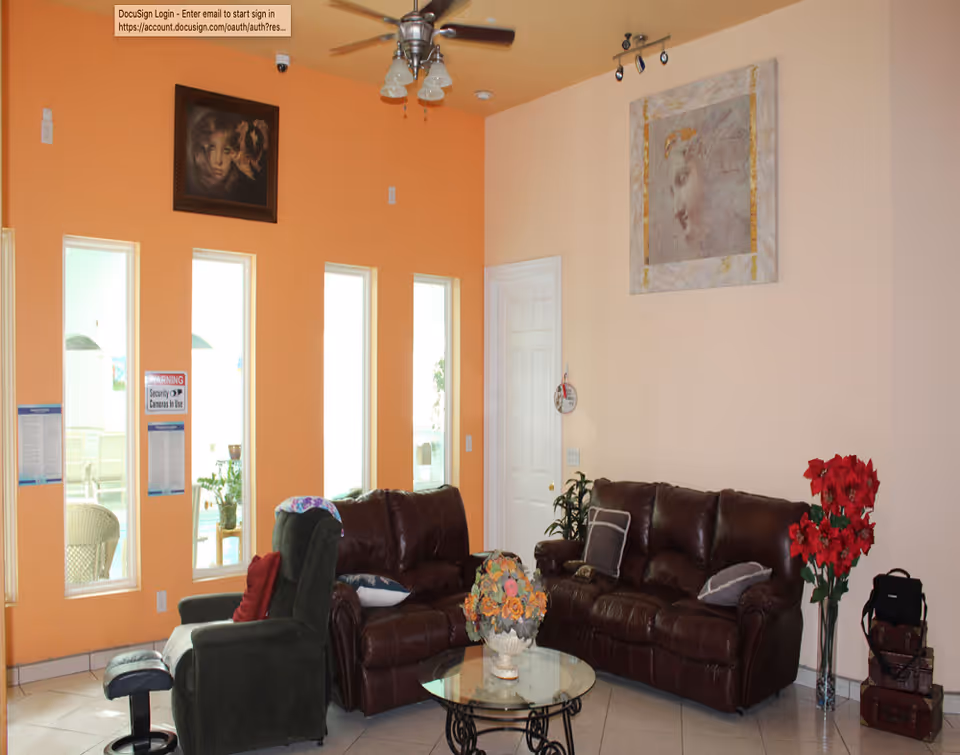 A living room with two brown leather sofas, a green recliner chair with an ottoman, a glass coffee table with a floral arrangement, and red flowers in a vase. The walls are painted orange and beige with two framed artworks. There are four tall windows letting in natural light and a ceiling fan with lights above.