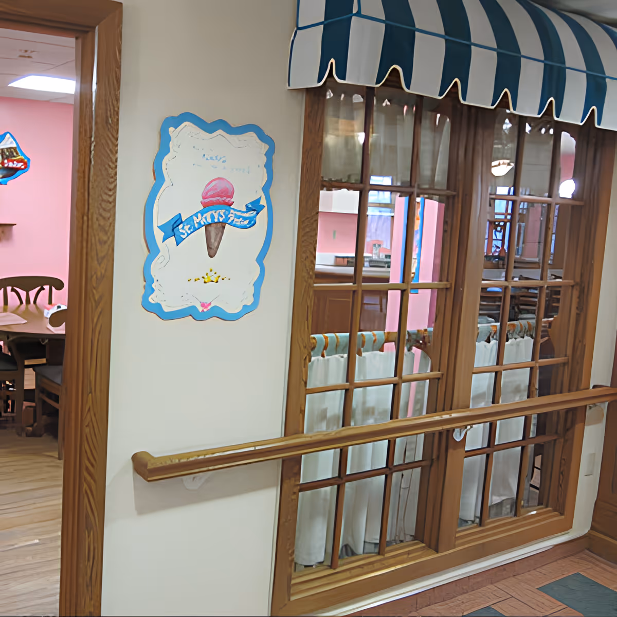 Interior view of a senior living facility hallway with wooden handrails and a window with blue and white striped awning. A sign on the wall features an ice cream cone with a blue ribbon that reads 'St. Mary's'. Through the window, a dining area with wooden chairs and pink walls is visible.