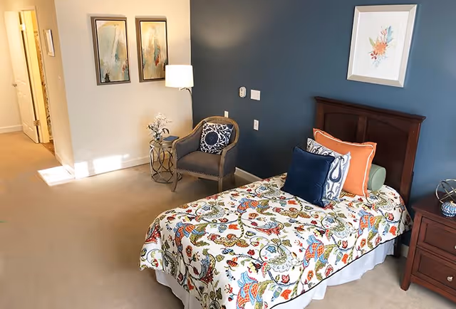 A cozy bedroom with a single bed covered in a colorful floral patterned bedspread and multiple decorative pillows. The room has a dark blue accent wall behind the bed, a wooden headboard, a wooden nightstand with a decorative item, and a comfortable armchair with a patterned cushion. Two framed abstract paintings hang on the adjacent light-colored wall, and a floor lamp provides soft lighting.