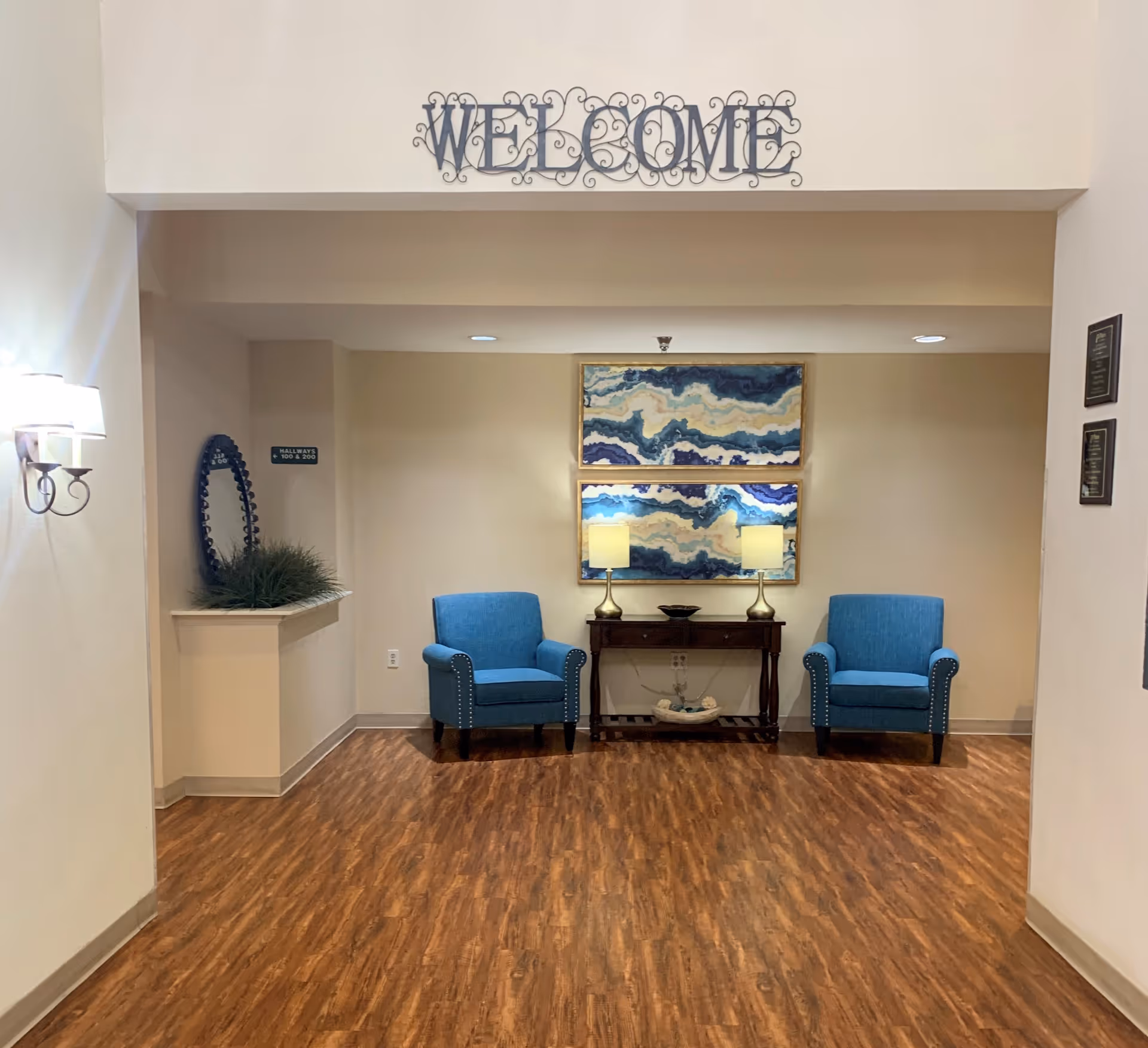 Lobby seating area with a 'WELCOME' sign above two blue armchairs, a central table with lamps, and abstract wall art.
