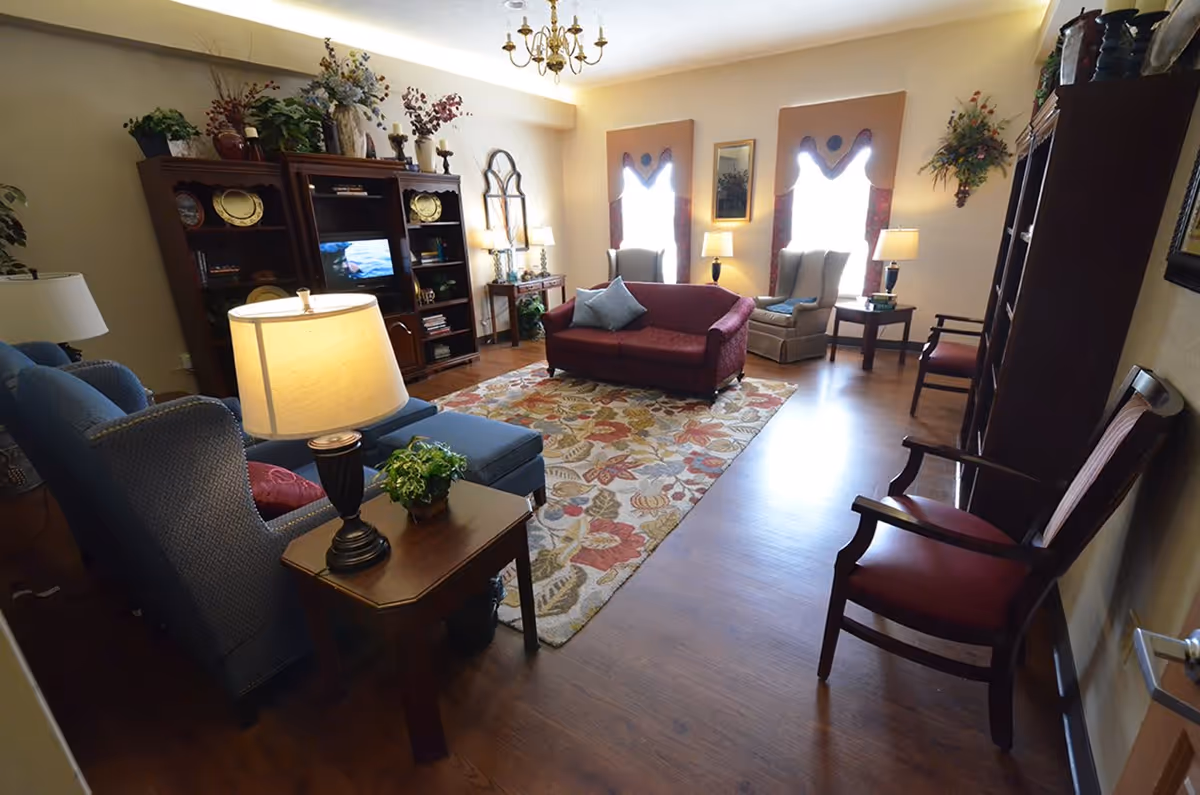 A cozy living room in a retirement center with a floral patterned rug, a red sofa, two beige armchairs near windows with decorative curtains, a blue armchair with an ottoman, wooden side tables with lamps, and a large wooden entertainment center with shelves and decorative items.