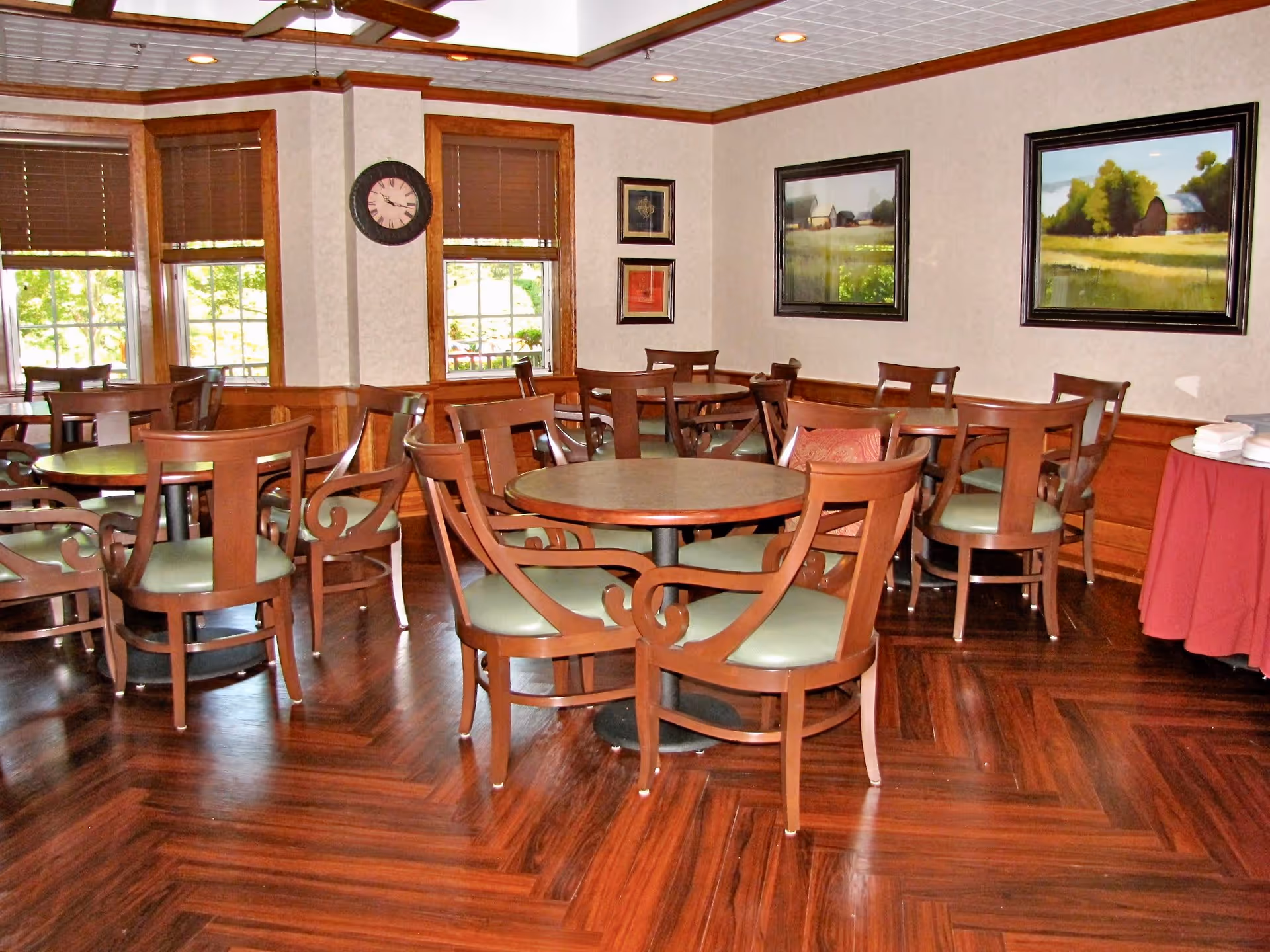 A dining room with multiple round wooden tables and matching chairs with green cushions. The room has wooden flooring, beige walls with wood paneling, several framed paintings, a clock on the wall, and windows with brown blinds letting in natural light. A table with a red tablecloth and stacked white plates is visible on the right side.