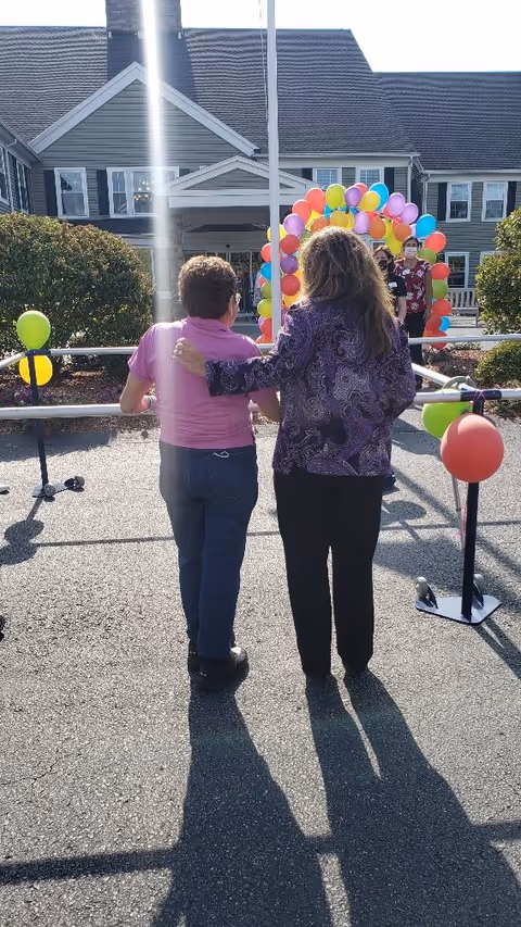 Two women standing outside in front of a building, one with her arm around the other. They are facing a colorful balloon arch and a person standing near it. The scene is sunny with shadows cast on the pavement.