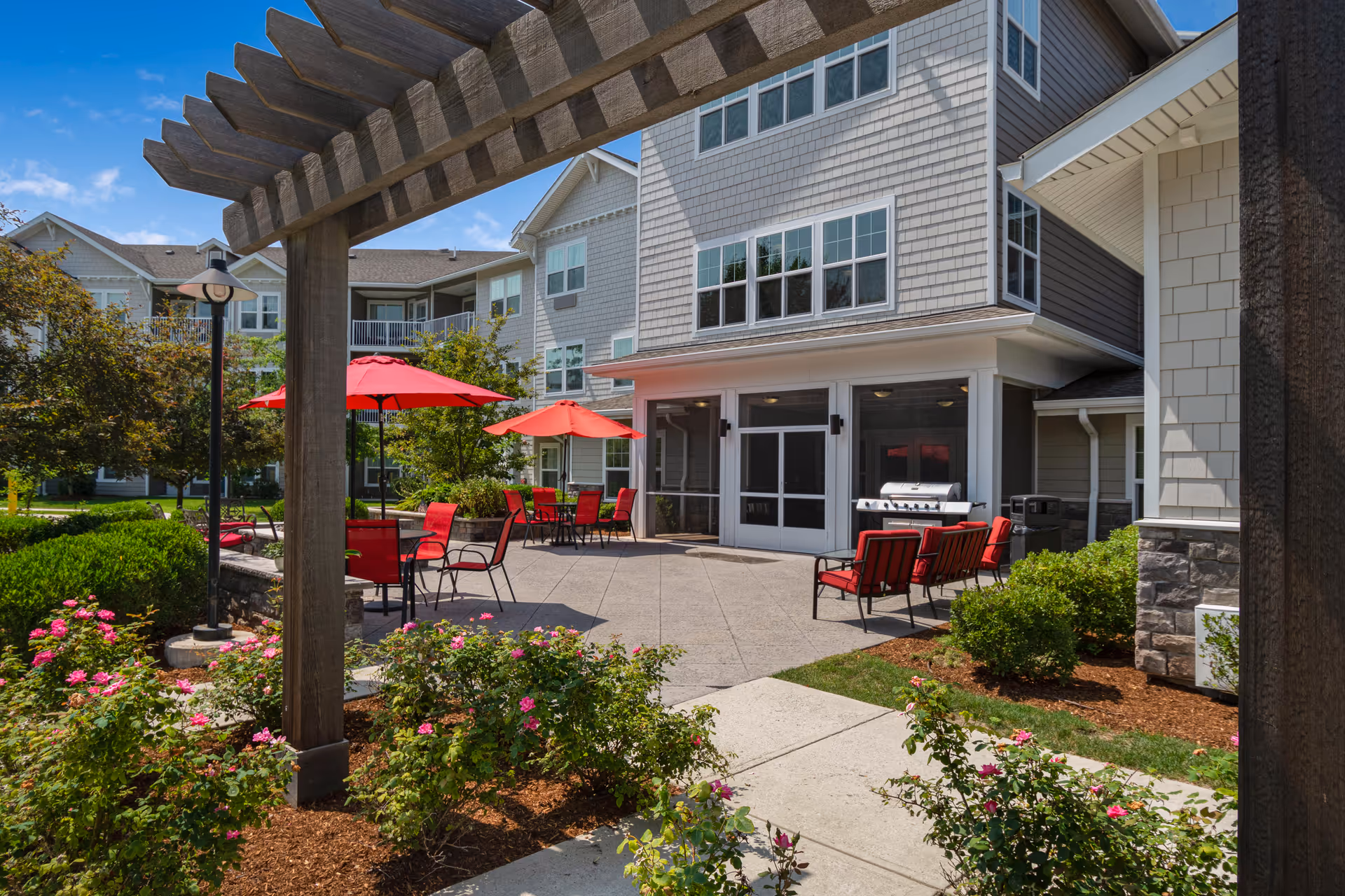 Outdoor patio area at Magnolia Heights Gracious Retirement Living featuring red patio chairs and tables with red umbrellas, a barbecue grill, landscaped flower beds with pink flowers, and a wooden pergola overhead. The building exterior is visible with multiple windows and light gray siding under a blue sky.