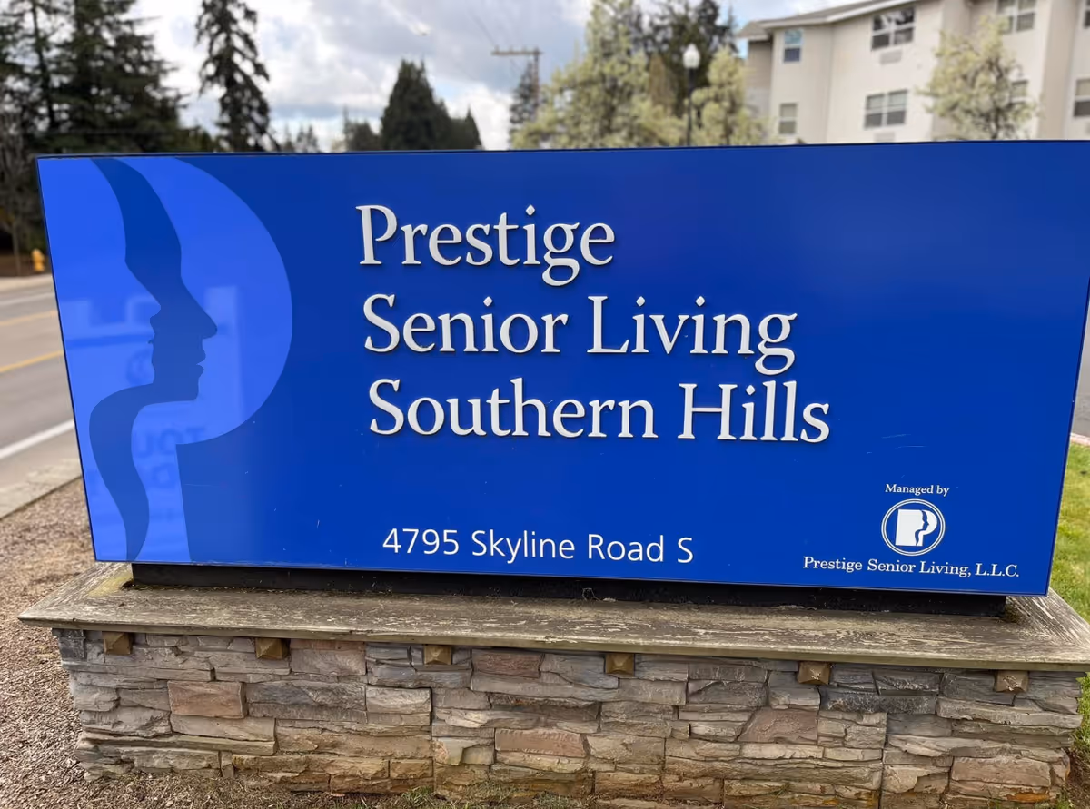 Blue roadside sign reading "Prestige Senior Living Southern Hills" with the address 4795 Skyline Road S mounted on a stone base in front of a building.