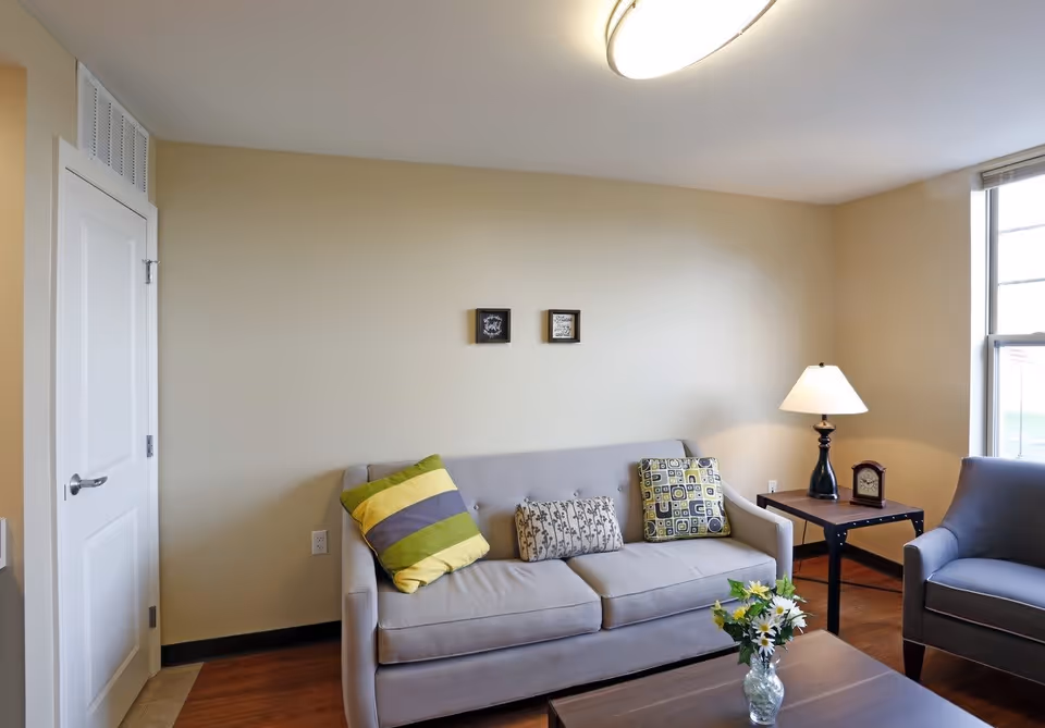 A cozy living room with a light gray sofa adorned with three decorative pillows in green, yellow, and patterned designs. Next to the sofa is a wooden side table with a black lamp and a small clock. A gray armchair is positioned near a window letting in natural light. A wooden coffee table with a vase of flowers is in the foreground. The walls are painted beige with two small framed pictures hanging above the sofa.