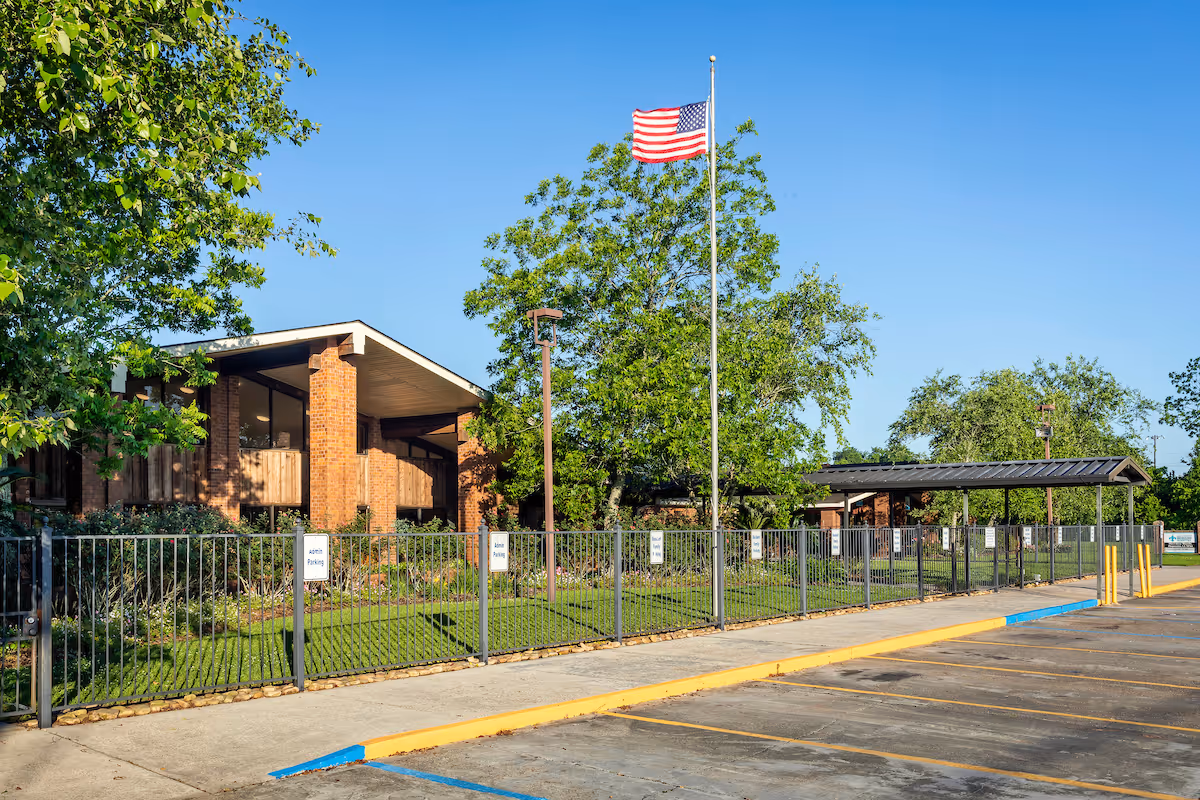 Exterior view of Legacy Nursing and Rehabilitation of Plaquemine showing a brick building with large windows, a fenced grassy area with trees, an American flag on a flagpole, and a parking lot in the foreground under a clear blue sky.
