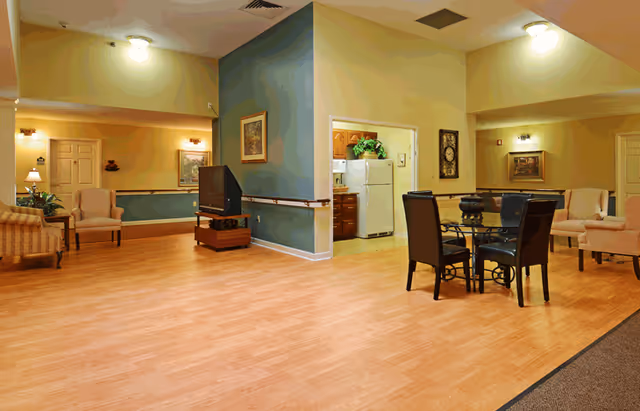 A spacious senior living common area with light wood flooring and soft yellow walls. The room features a small dining table with four chairs, several armchairs, a TV on a stand, framed artwork on the walls, and a view into a kitchen area with a white refrigerator and wooden cabinets. The space is well-lit with ceiling and wall lights.
