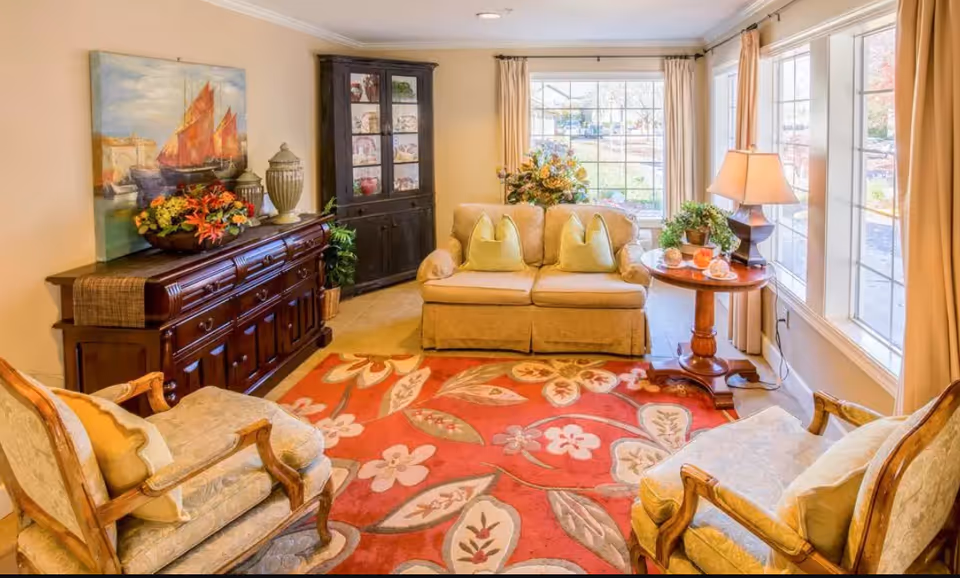 Bright living room with a loveseat, two upholstered armchairs, a red floral rug, wooden sideboard and large windows letting in daylight.