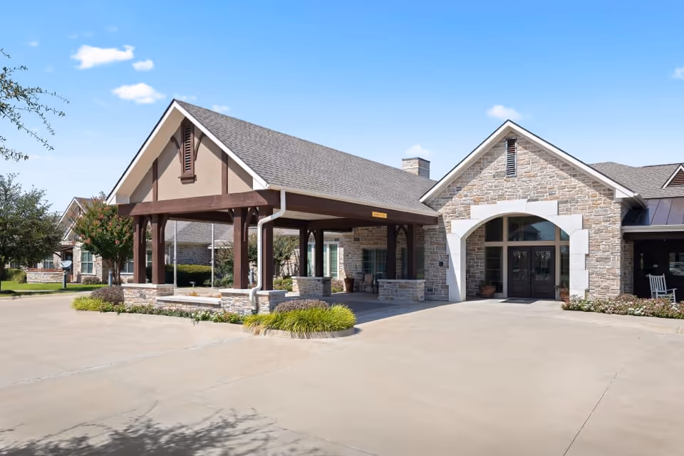 Exterior view of a senior living facility named Village on the Park Stonebridge Ranch featuring a covered entrance with wooden beams, stone walls, and a driveway under a clear blue sky.