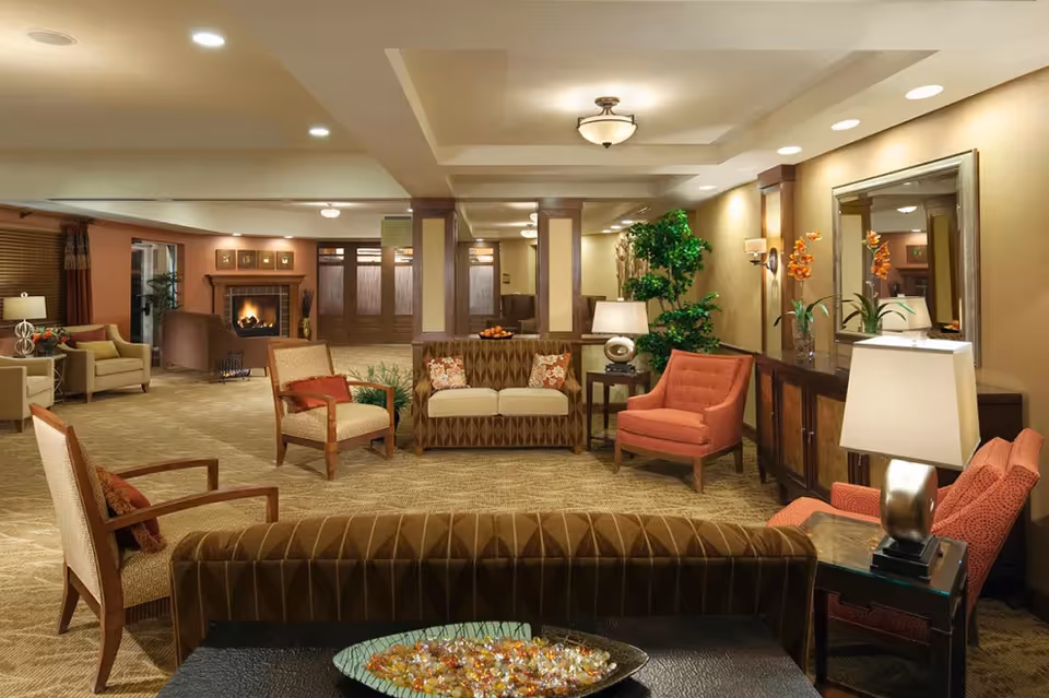 A spacious and warmly lit living room area in an assisted living facility featuring multiple upholstered chairs and sofas arranged around a central coffee table. The room has beige walls, carpeted floors, a fireplace in the background, decorative plants, and lamps providing soft lighting. There is a large mirror on one wall and a sideboard with flower arrangements.