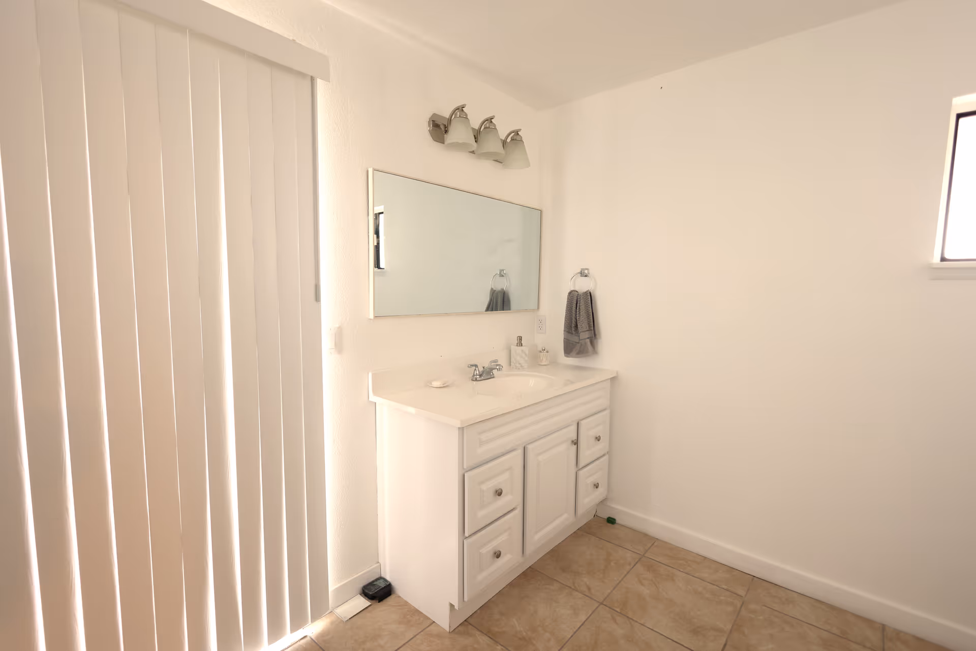 A clean bathroom with a white vanity cabinet, a sink, a large rectangular mirror above it, a three-light fixture, a gray hand towel hanging on a ring, beige tiled floor, and vertical blinds covering a window or door on the left side.