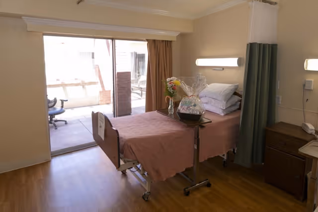 A hospital-style room with a single bed covered in a pink blanket, a bedside table with a telephone, and a small table with a gift basket and flowers. The room has wooden flooring, a sliding glass door leading to an outdoor patio, and a privacy curtain beside the bed.