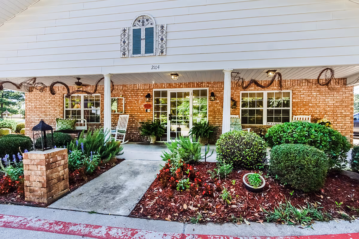 Front entrance of a brick building with a covered porch featuring white columns, rocking chairs, potted plants, and decorative greenery. The walkway is flanked by landscaped flower beds with bushes and flowers. The building number 2104 is visible above the porch.