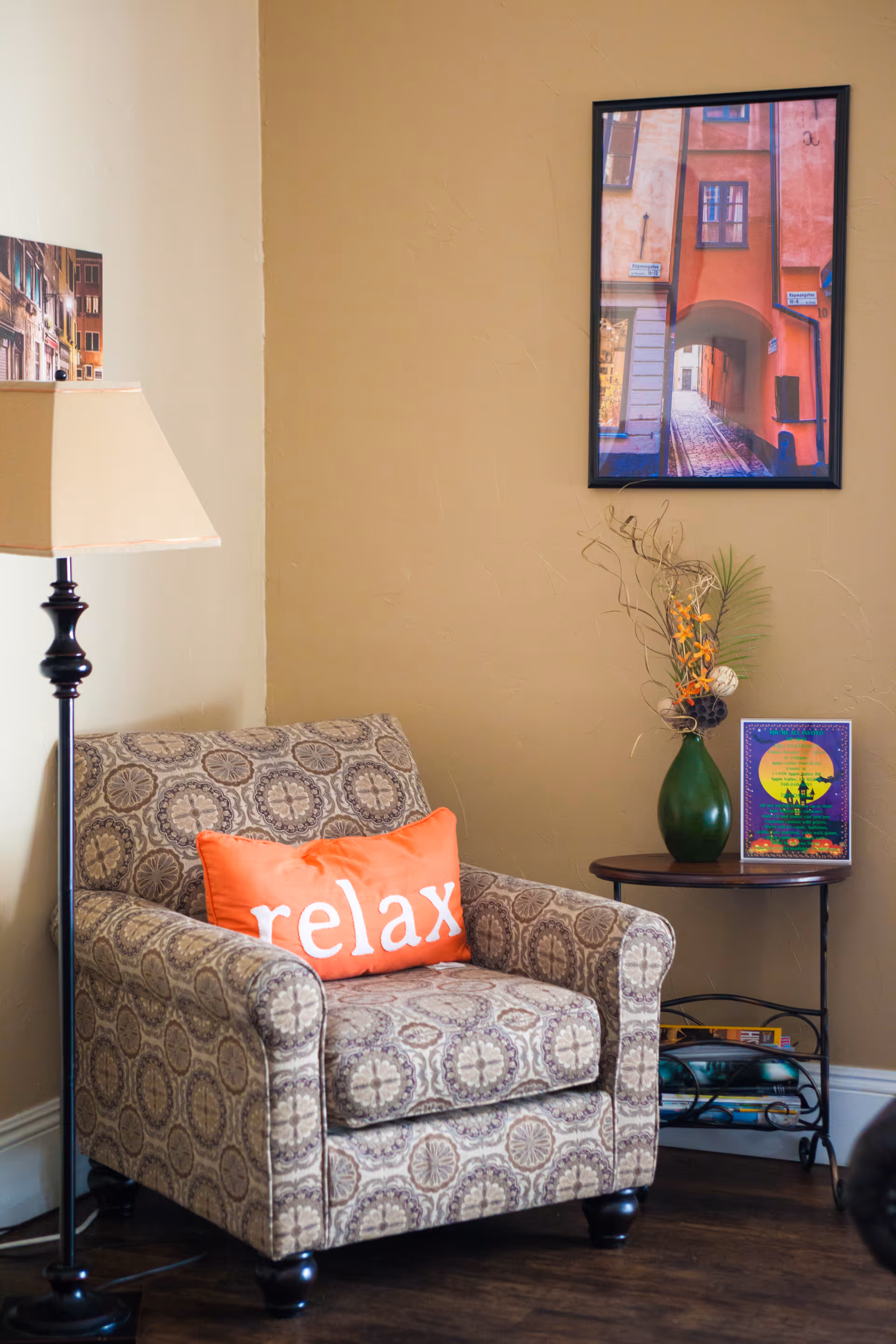A cozy corner with a patterned armchair featuring an orange pillow with the word 'relax' on it. Next to the chair is a floor lamp with a beige shade and a small round table holding a green vase with decorative branches and a framed picture. A framed photo of a narrow street is hung on the beige wall behind the chair.