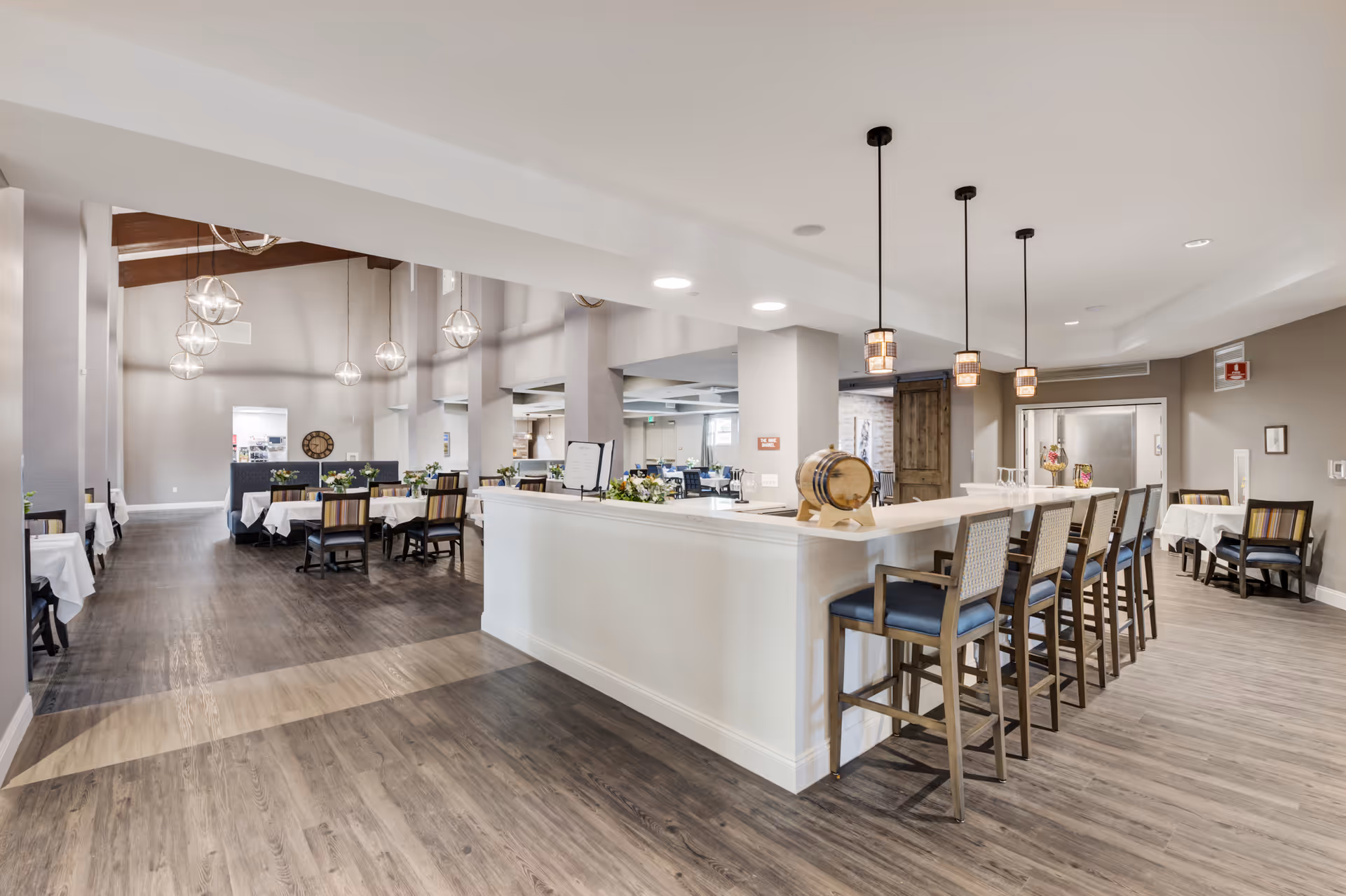 Bright open dining and lounge area with a long white bar counter and stools, multiple dining tables, and pendant lighting.