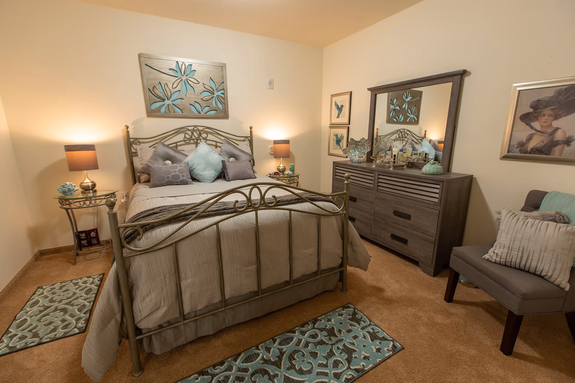 A cozy bedroom with a metal frame bed adorned with multiple pillows and a beige comforter. On either side of the bed are matching glass-top nightstands with lamps. A large wooden dresser with a mirror is against the wall, decorated with various items. The room also features a cushioned chair with pillows, framed artwork on the walls, and patterned rugs on the carpeted floor.
