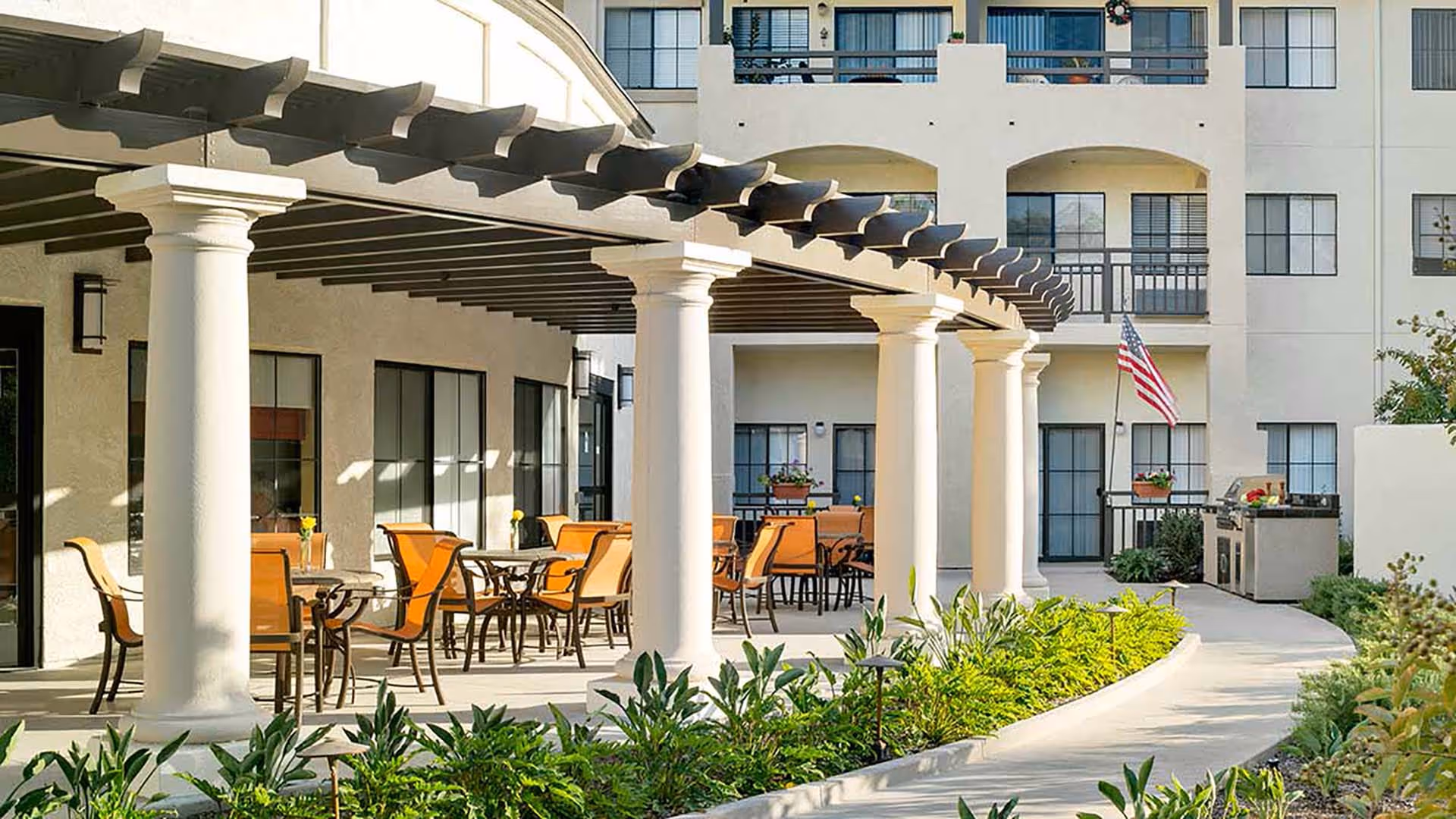 Outdoor patio area at Atria Hillcrest featuring a covered seating space with multiple tables and orange chairs, white columns supporting a pergola, a walkway surrounded by greenery, and an American flag near a barbecue grill.
