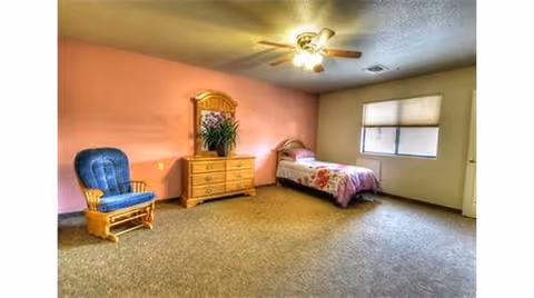 A simple bedroom with a single bed covered in a floral bedspread, a wooden dresser with a mirror and a plant on top, a blue cushioned rocking chair, a ceiling fan with lights, and a window with blinds partially closed. The walls are painted pink and beige, and the floor is carpeted.