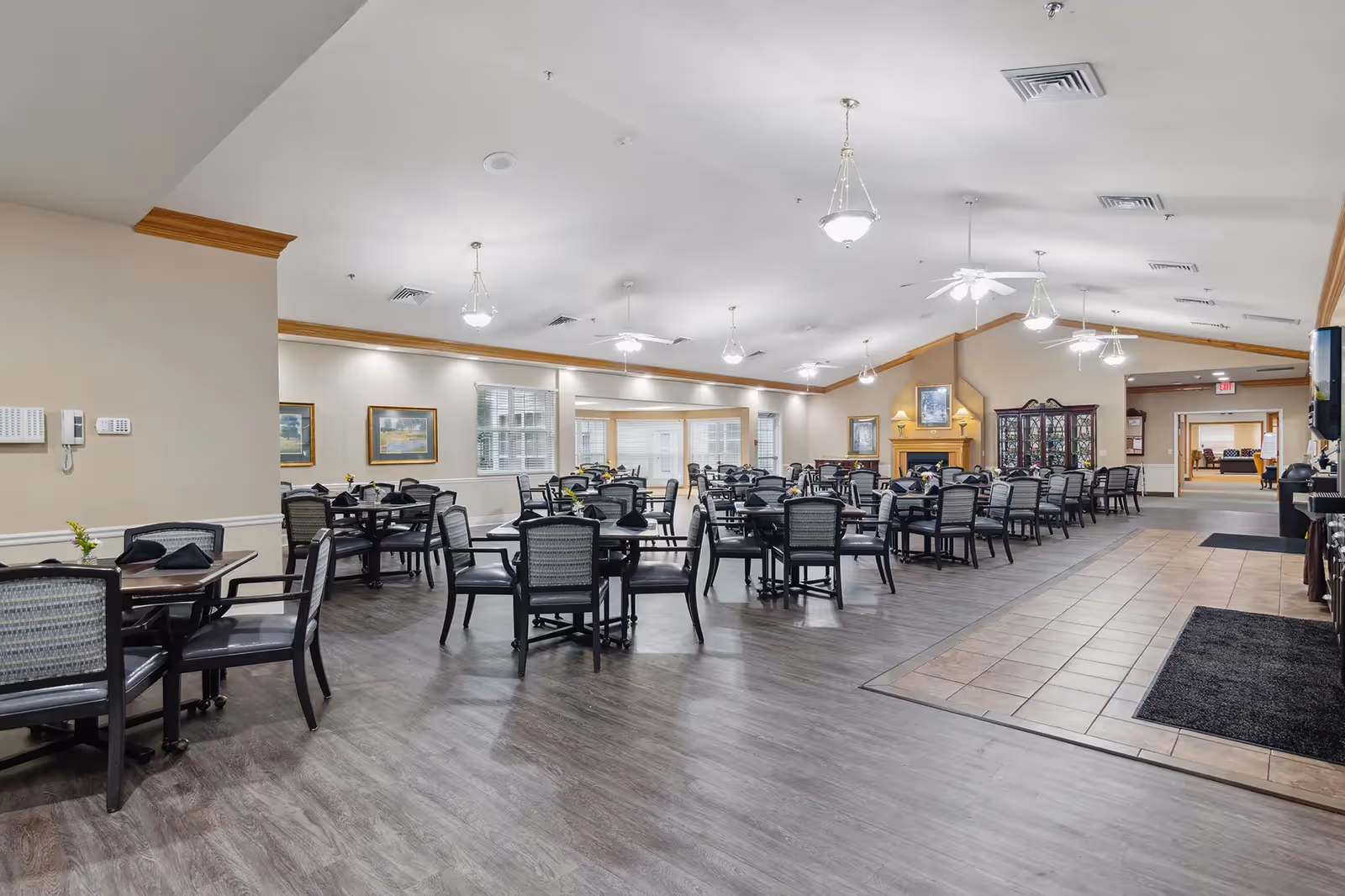 A spacious dining room with multiple tables and chairs arranged neatly. The room features a combination of wood and tile flooring, ceiling fans, pendant lights, framed artwork on the walls, and a fireplace with a cabinet nearby. The area is well-lit with natural light coming through the windows.